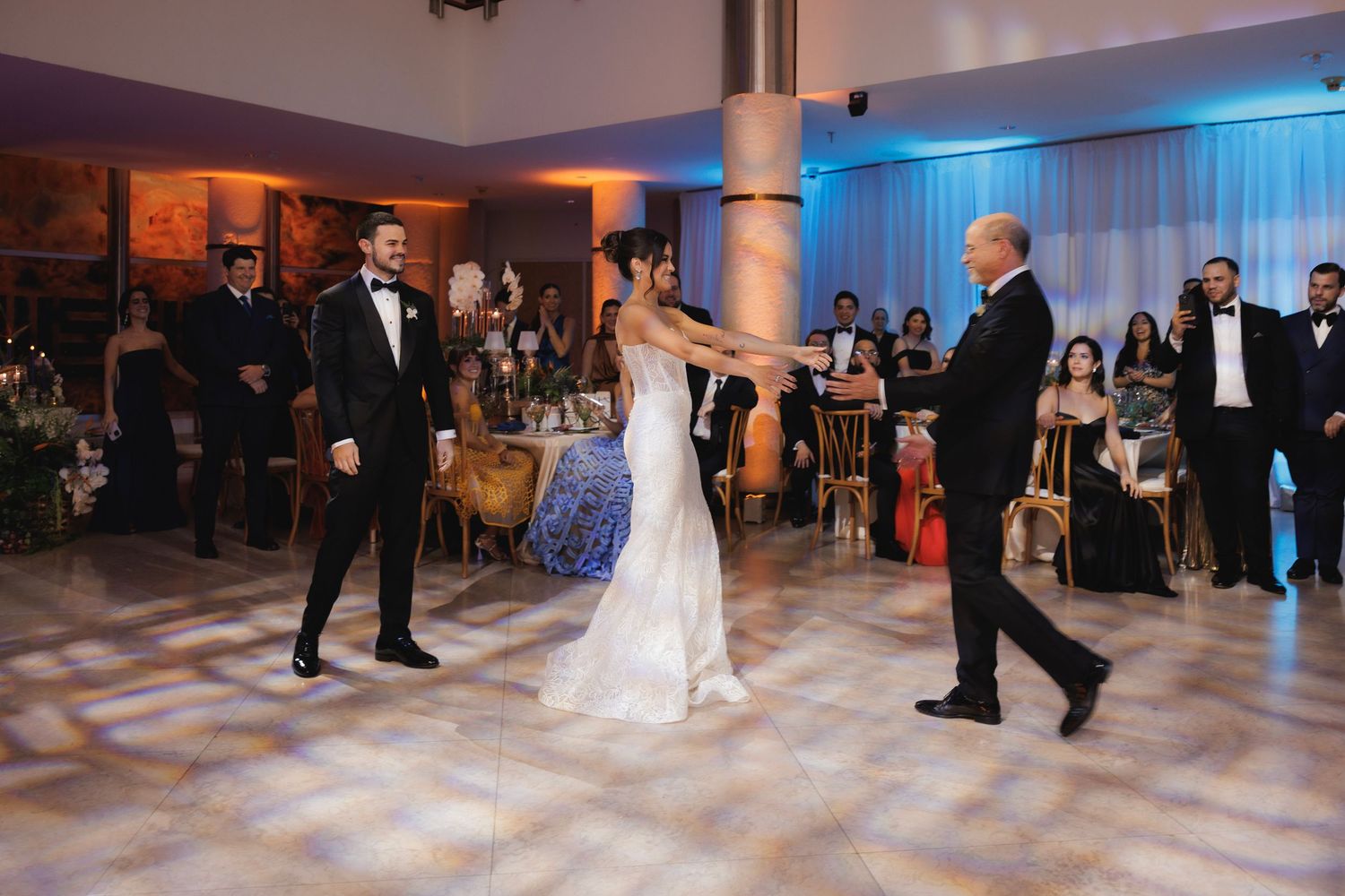Elegant wedding dance party in a spacious ballroom with blue mood lighting and decorative columns.
