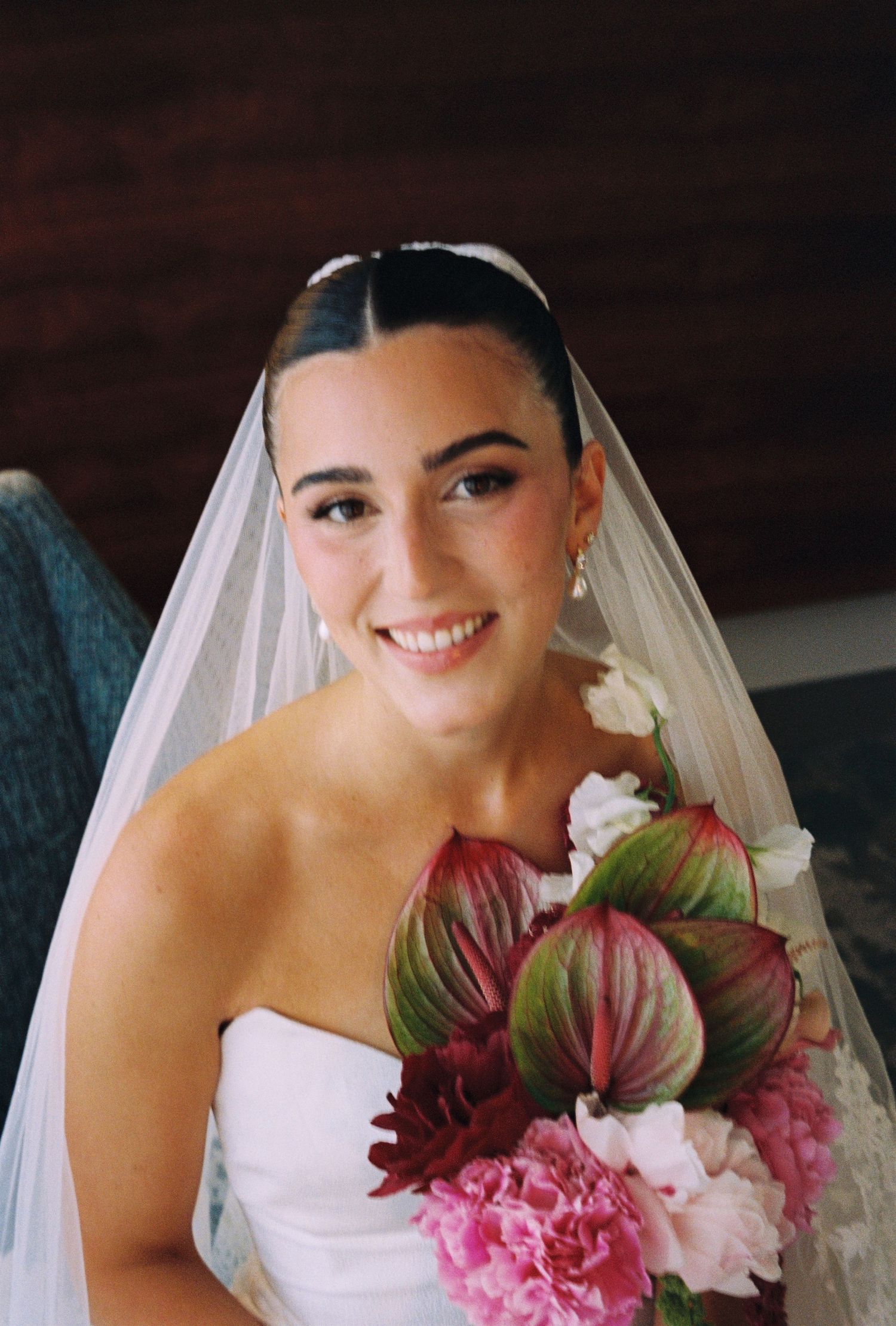 Smiling bride in white strapless gown and veil holding a bouquet of pink and burgundy flowers.