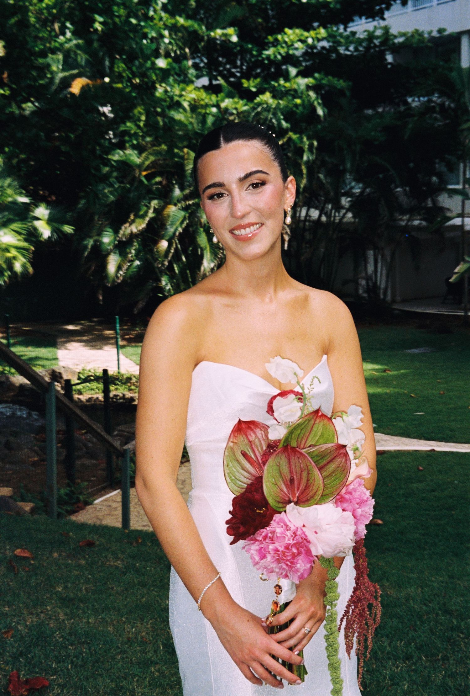 Smiling bride in strapless white dress holding a vibrant pink and red floral bouquet outdoors in sunlight.
