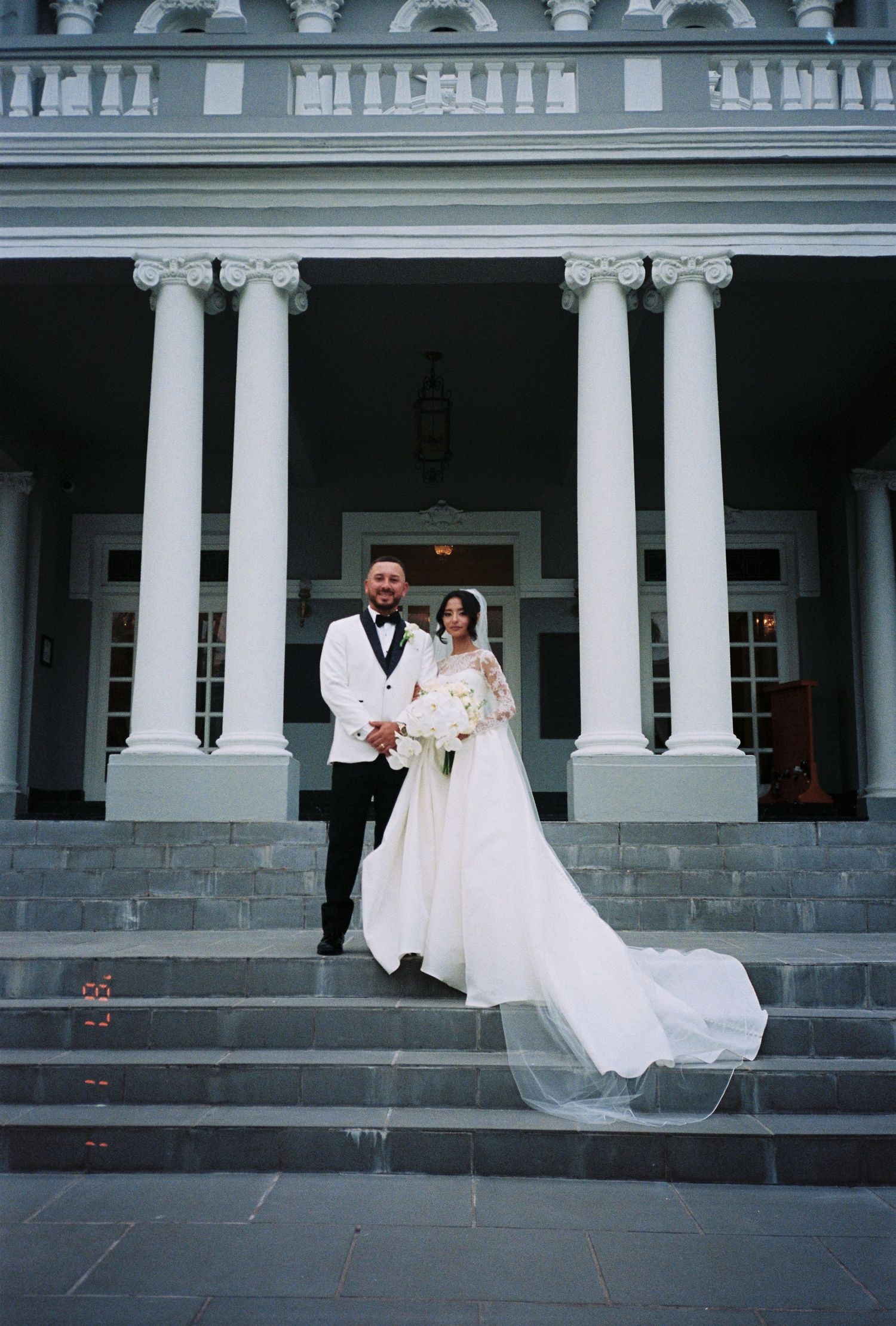 Bride and groom posing on grand white columned mansion steps, bride in elegant gown with long veil, groom in white tuxedo.
