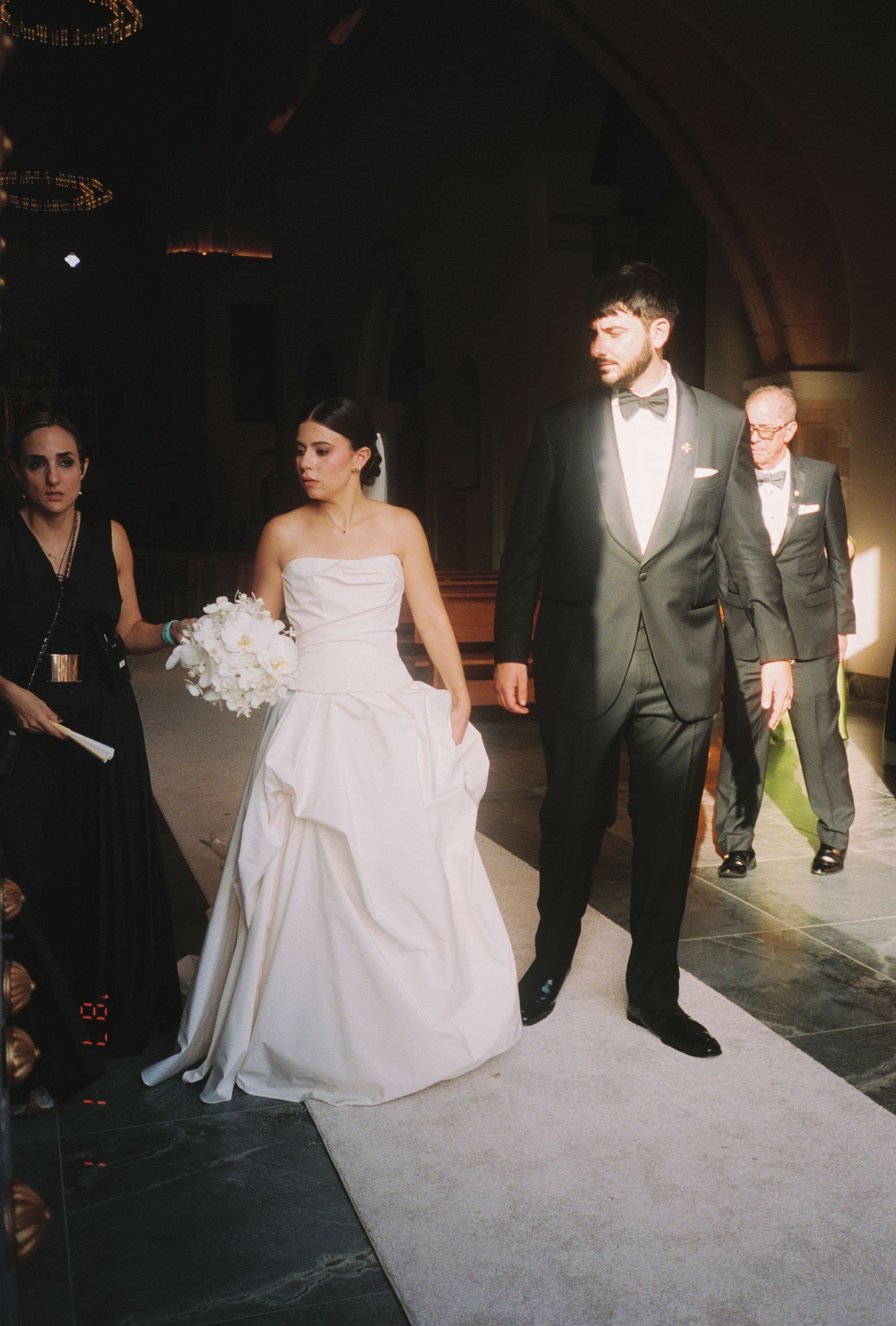 Bride in strapless white gown and groom in dark tuxedo walk down aisle after wedding ceremony.