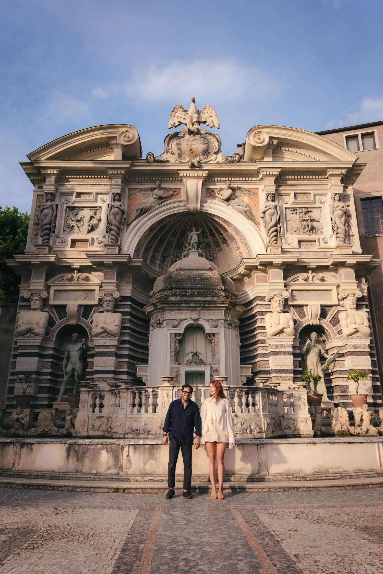 Couple posing in front of ornate baroque fountain with eagle sculpture and classical statues in Tivoli, Italy.