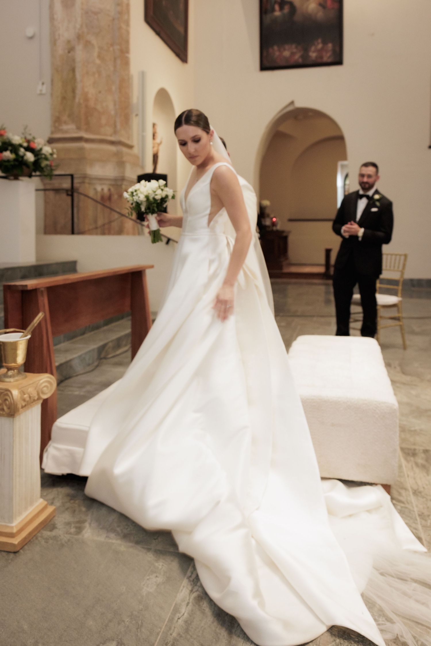 A bride in a flowing white gown walks through an elegant church with stone columns.