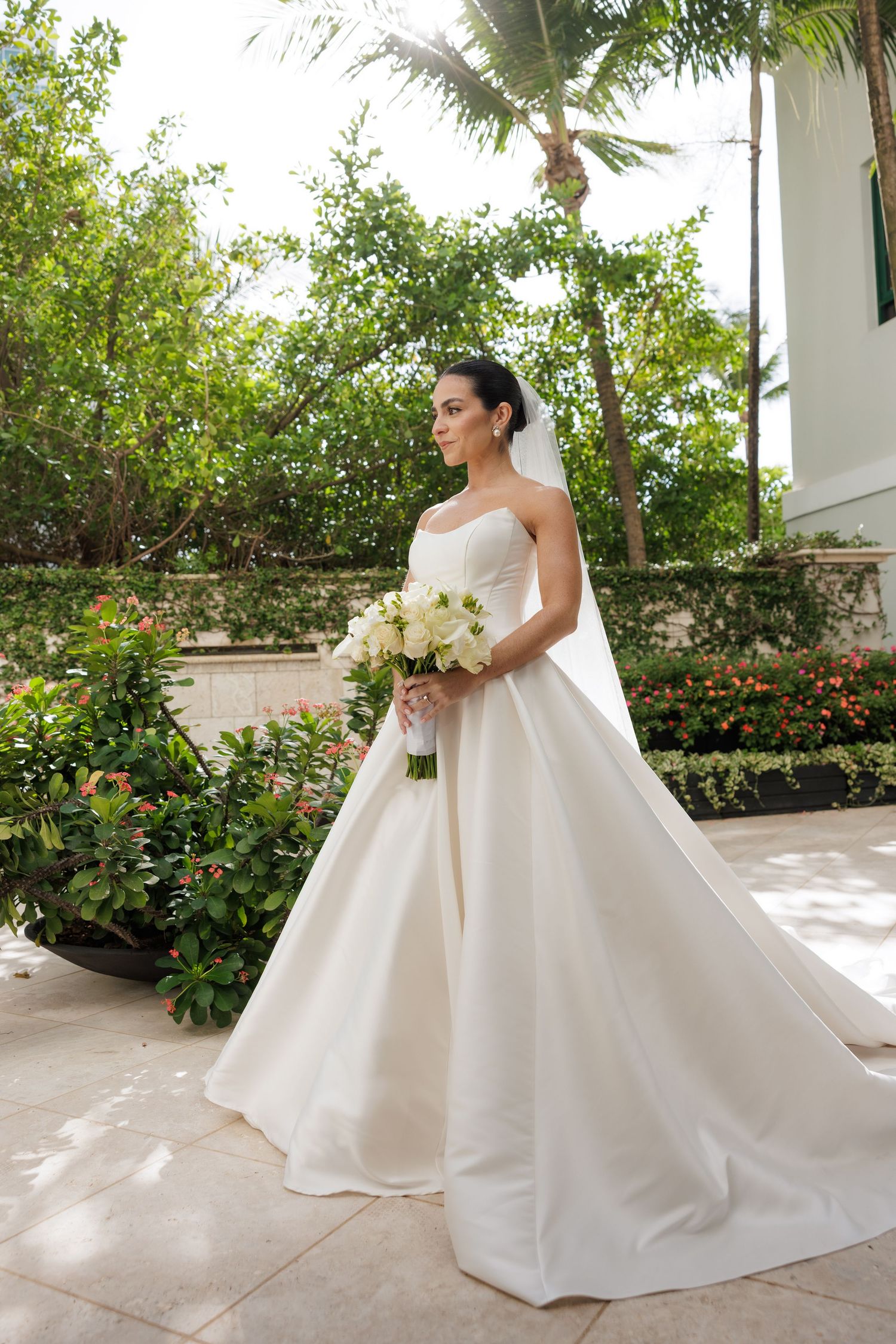 Bride in flowing white gown holds bouquet while posing in outdoor garden setting.