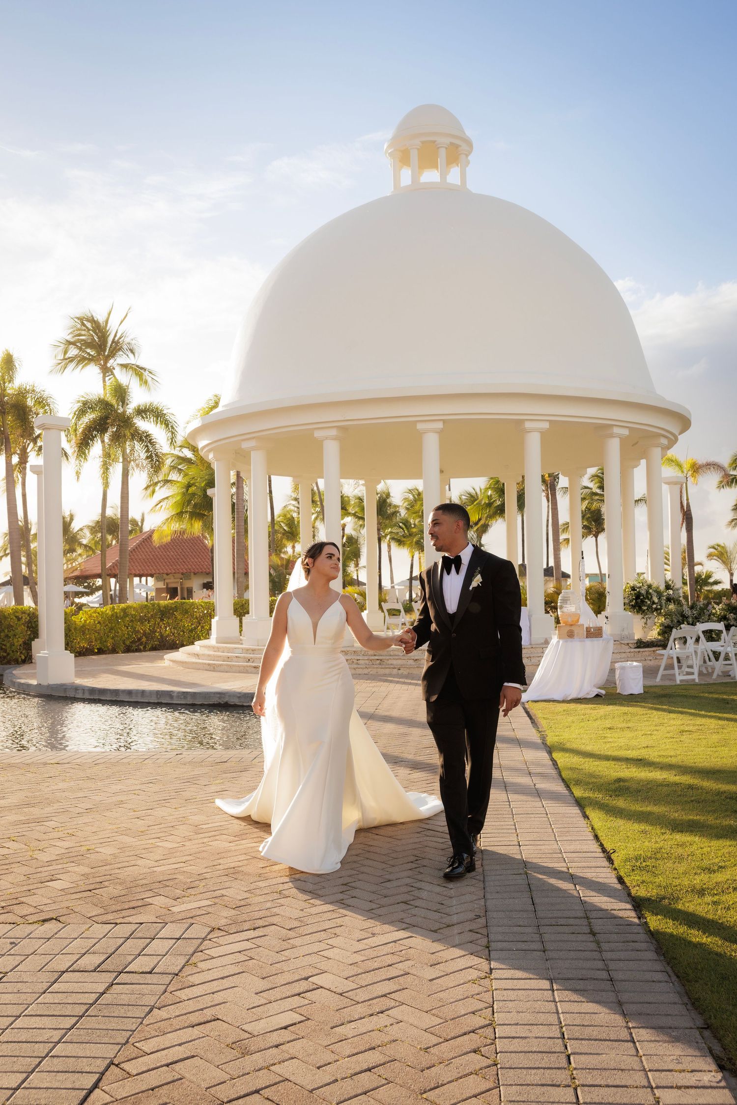 Newlyweds walk along path by elegant white domed gazebo with palm trees and beautiful sunset lighting at tropical venue.