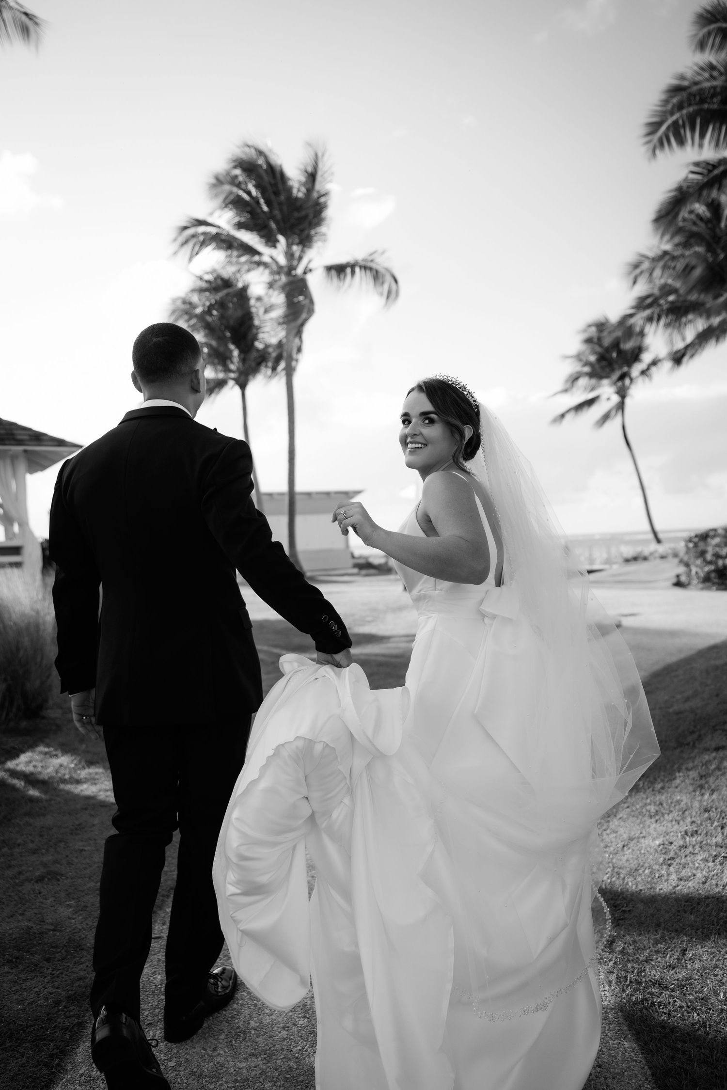 A bride and groom share a playful moment in the wind with palm trees swaying in the background.