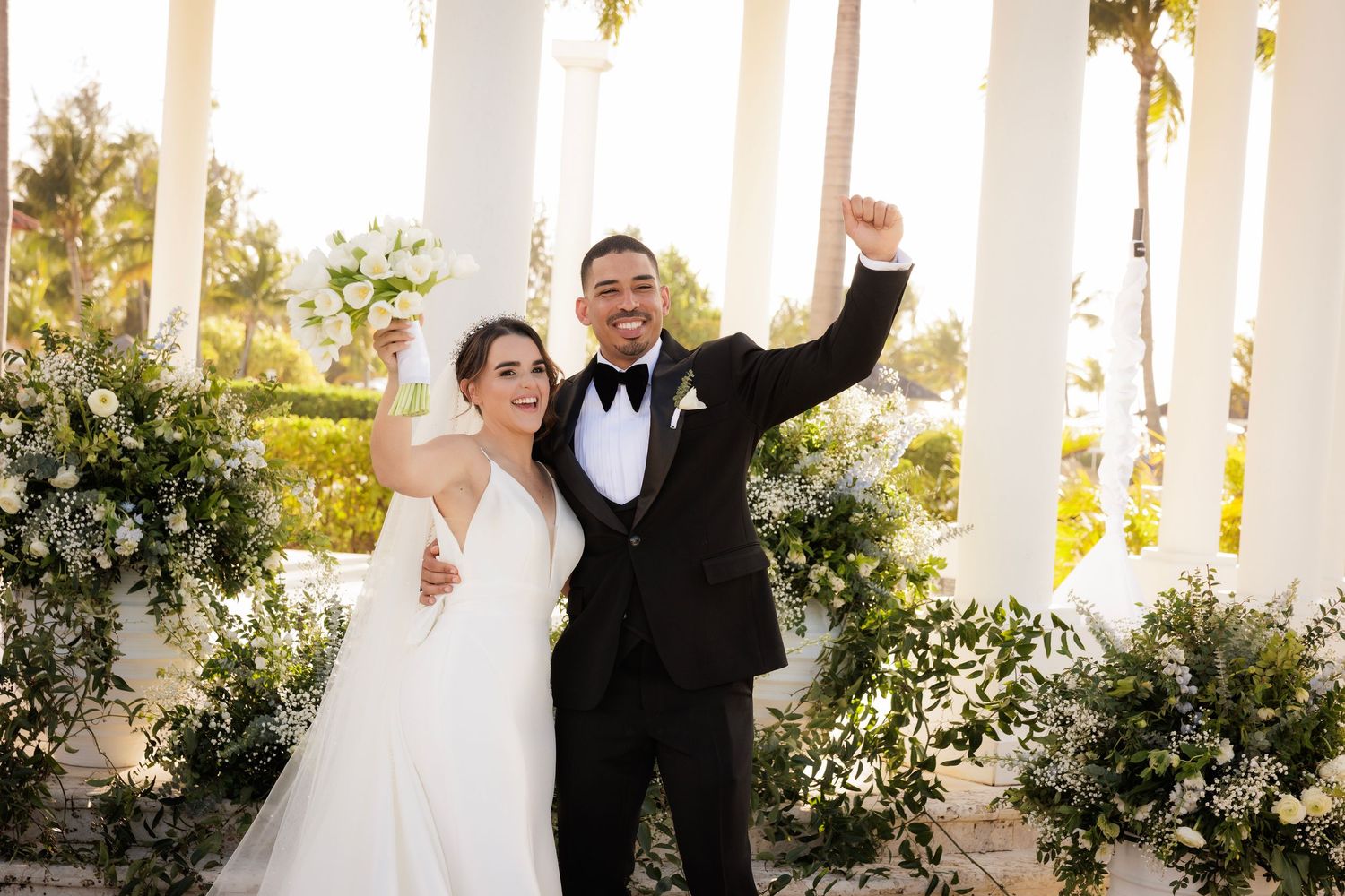 Wedding couple poses joyfully under white outdoor columns decorated with green foliage during sunset.