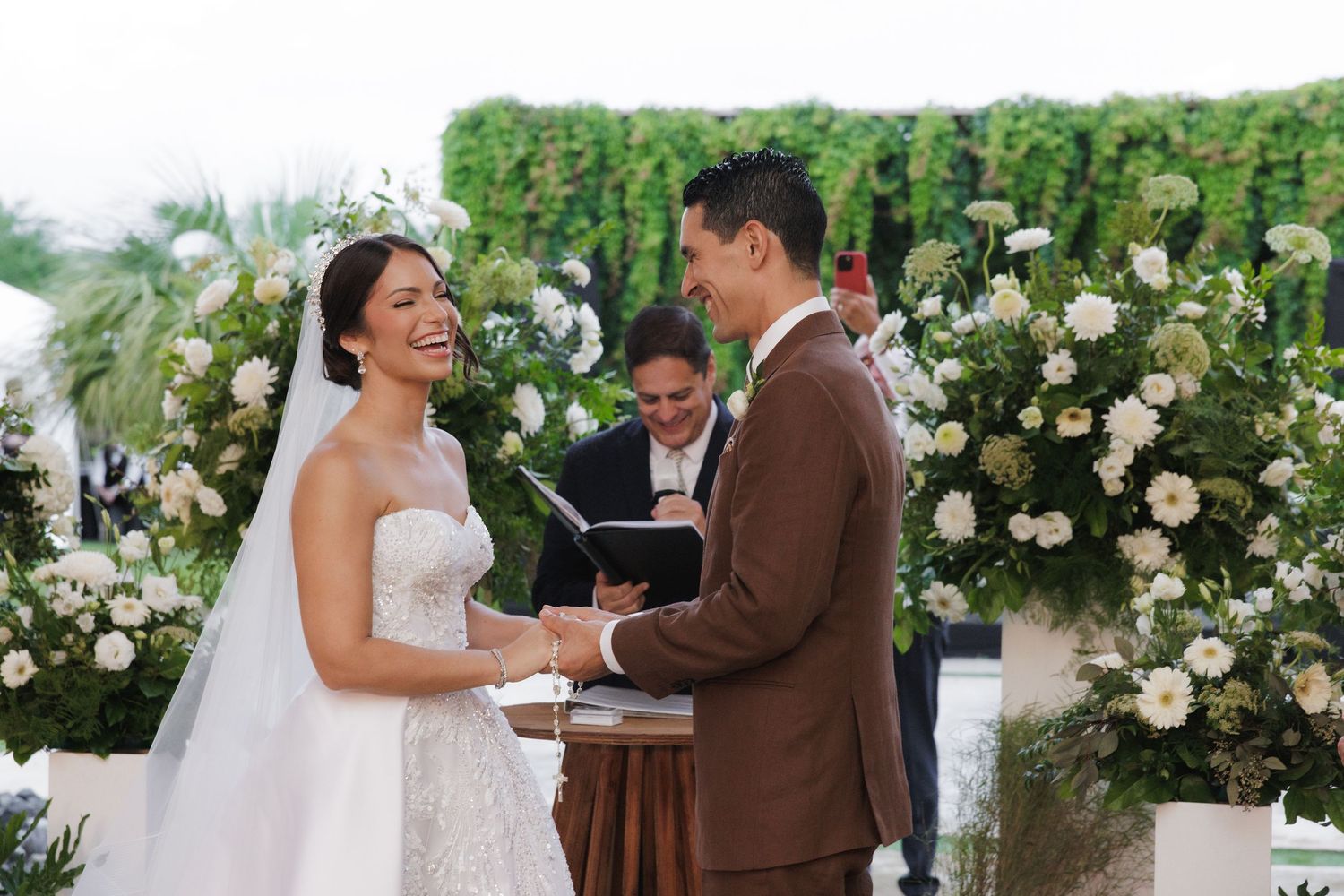 A joyful outdoor wedding ceremony takes place under a lush floral arch with white roses and greenery.