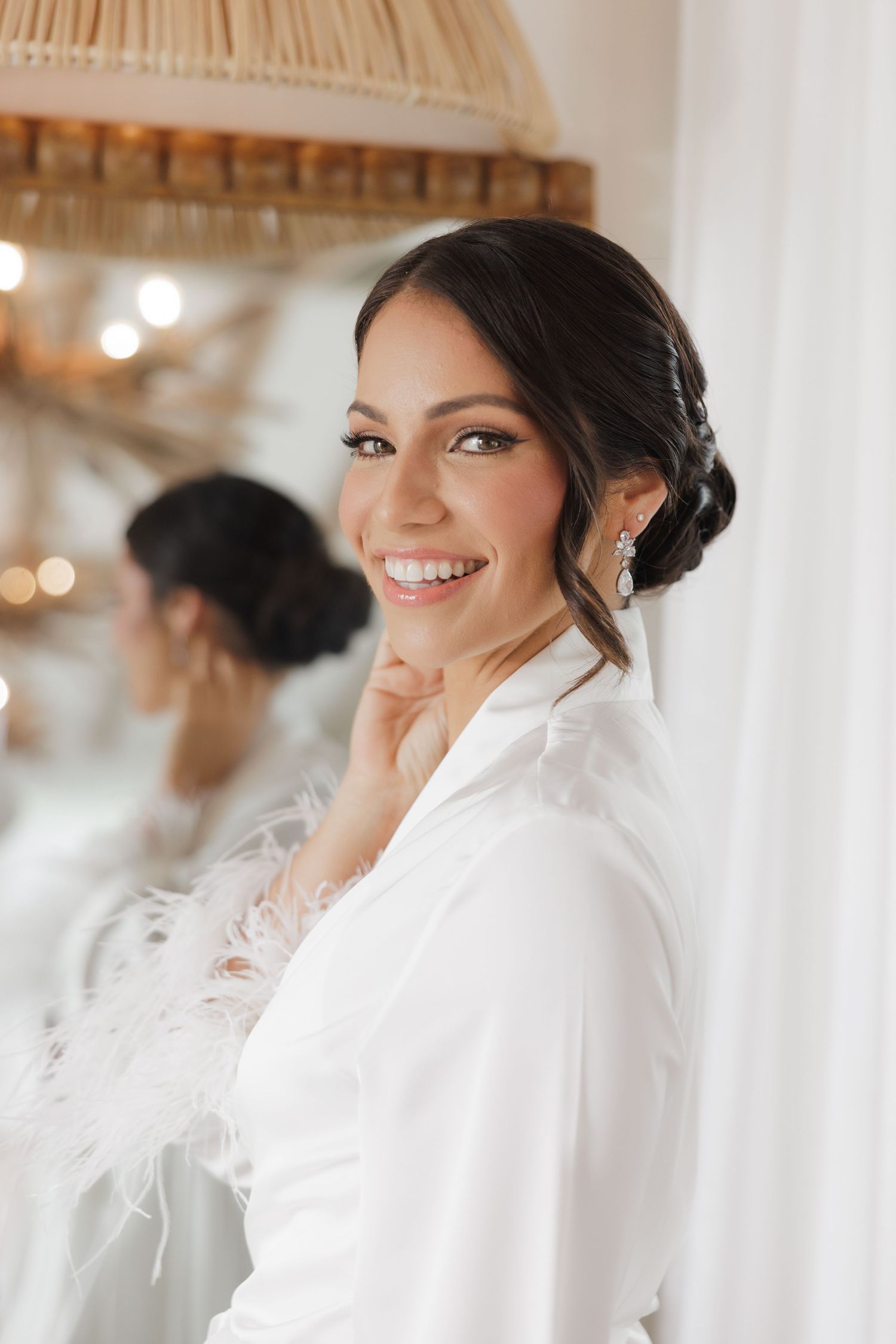 A bride getting ready in a white feather-trimmed robe smiles while preparing for her wedding day.