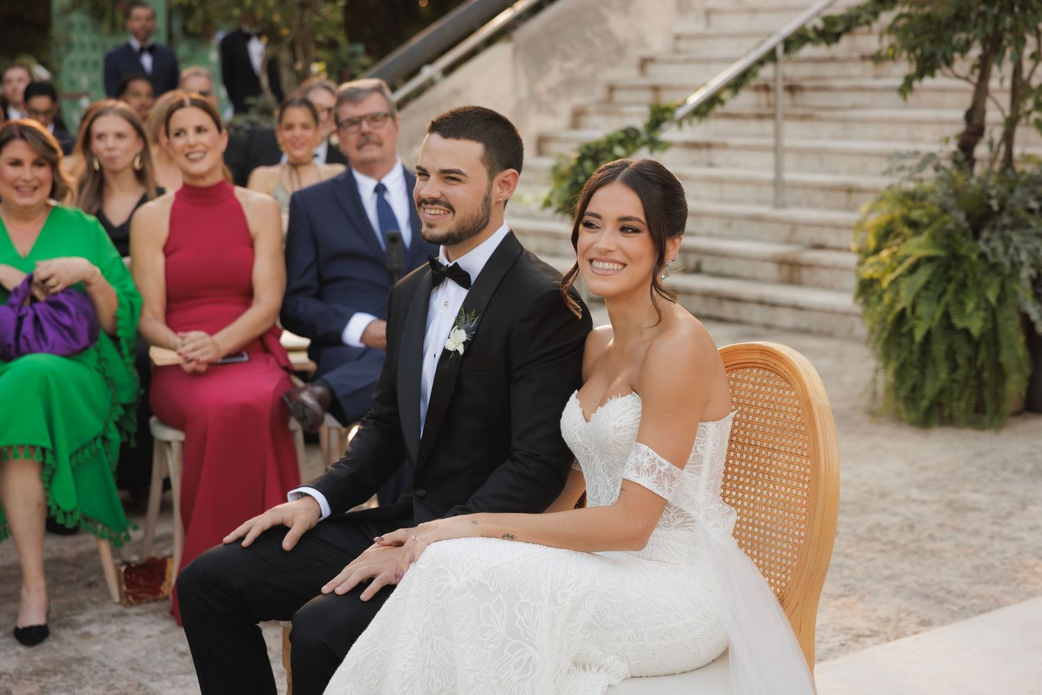 Wedding attendees stand on stone steps during an outdoor ceremony with decorative greenery and white fabric.