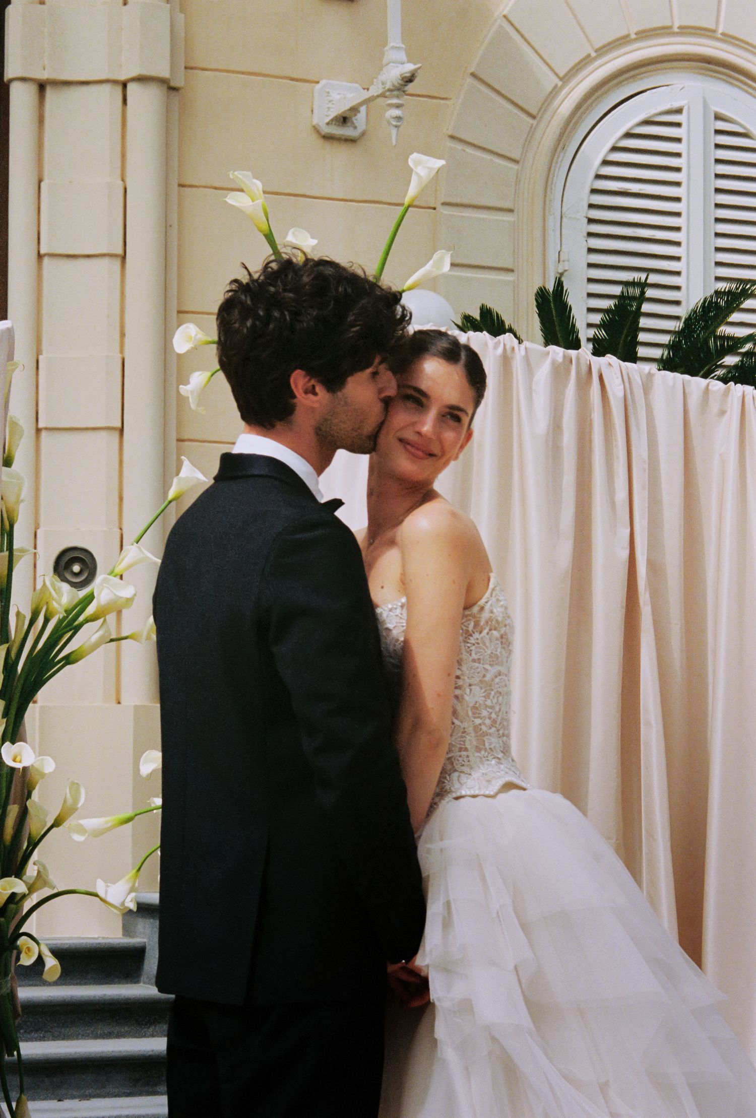 Bride and groom share a romantic kiss at an outdoor wedding ceremony with white calla lilies and draped fabric backdrop.