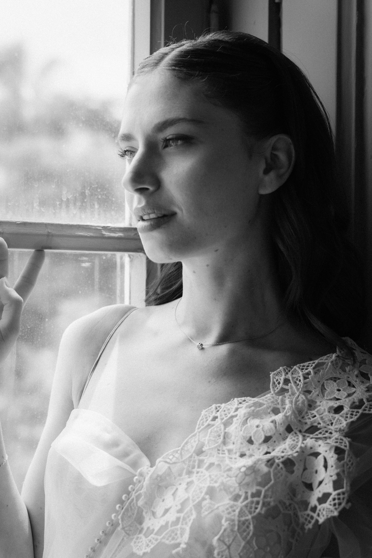 Black and white portrait of a young woman in lace dress gazing thoughtfully out a rain-speckled window.