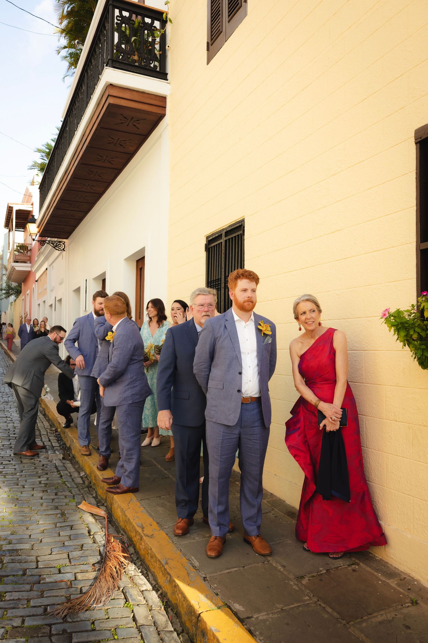Wedding guests line up along a yellow wall on a cobblestone path during a formal event.