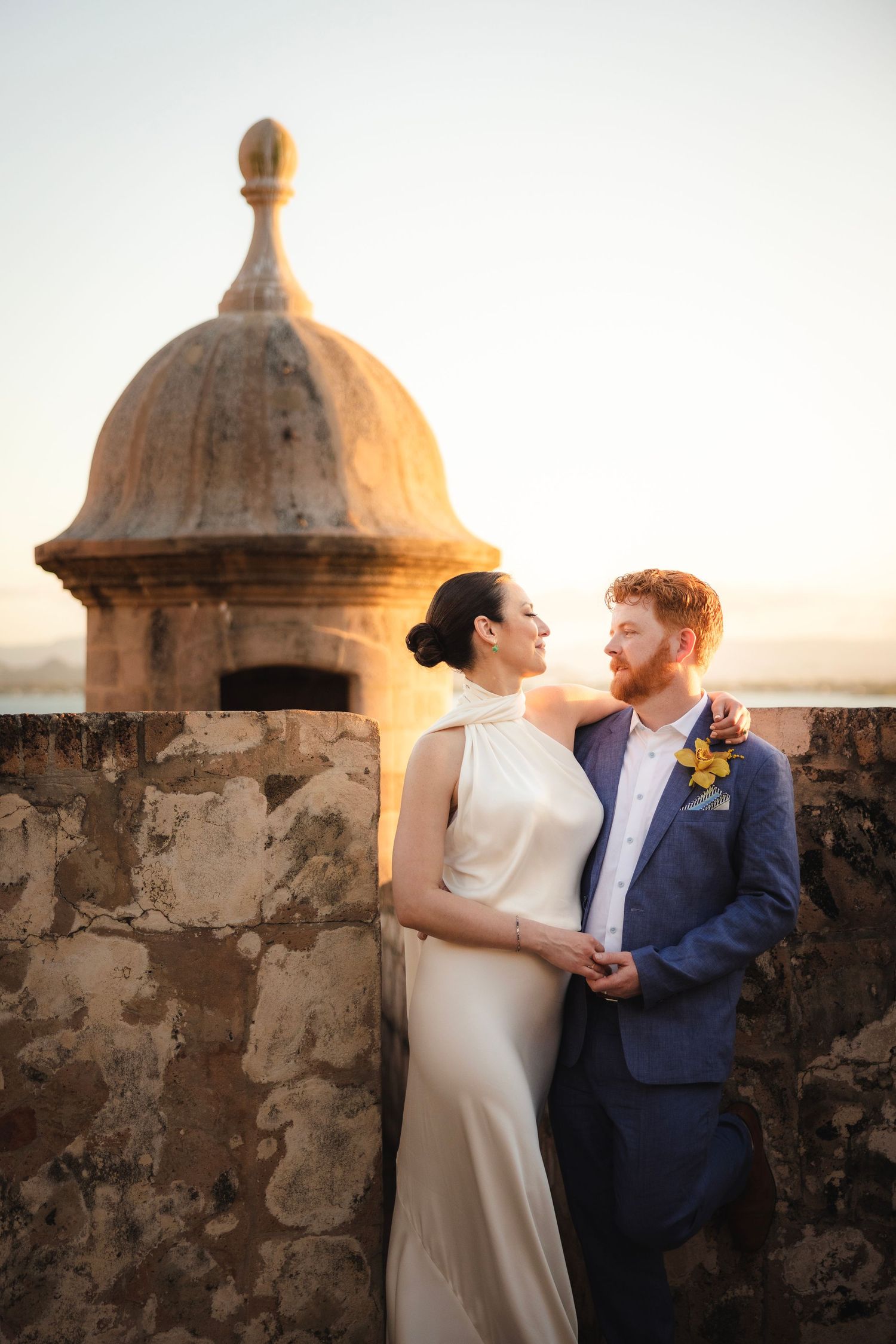 A couple in wedding attire shares a romantic moment at sunset near a historic stone watchtower with domed architecture.