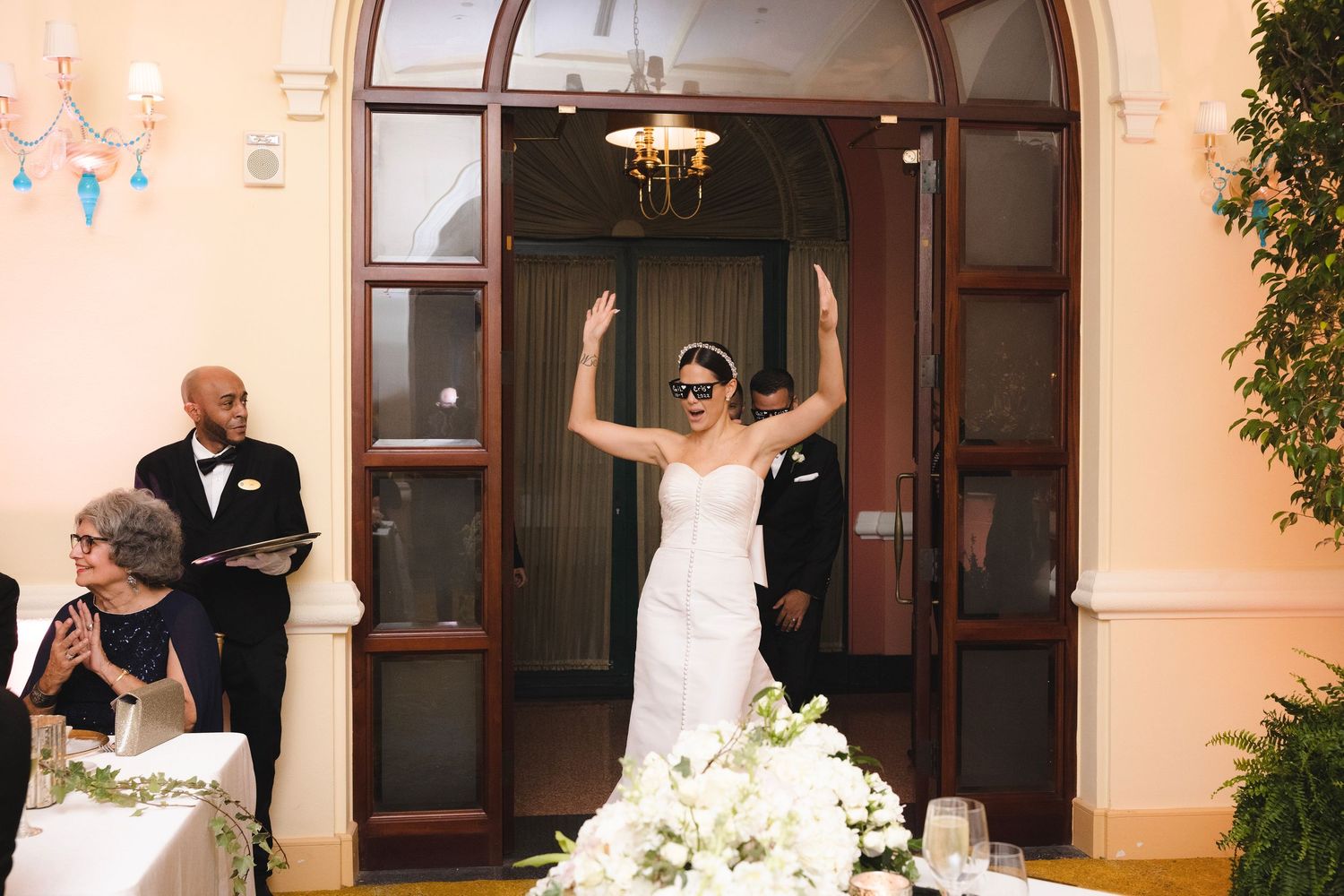 A joyful wedding celebration outside ornate wooden doors with white floral decorations at sunset.