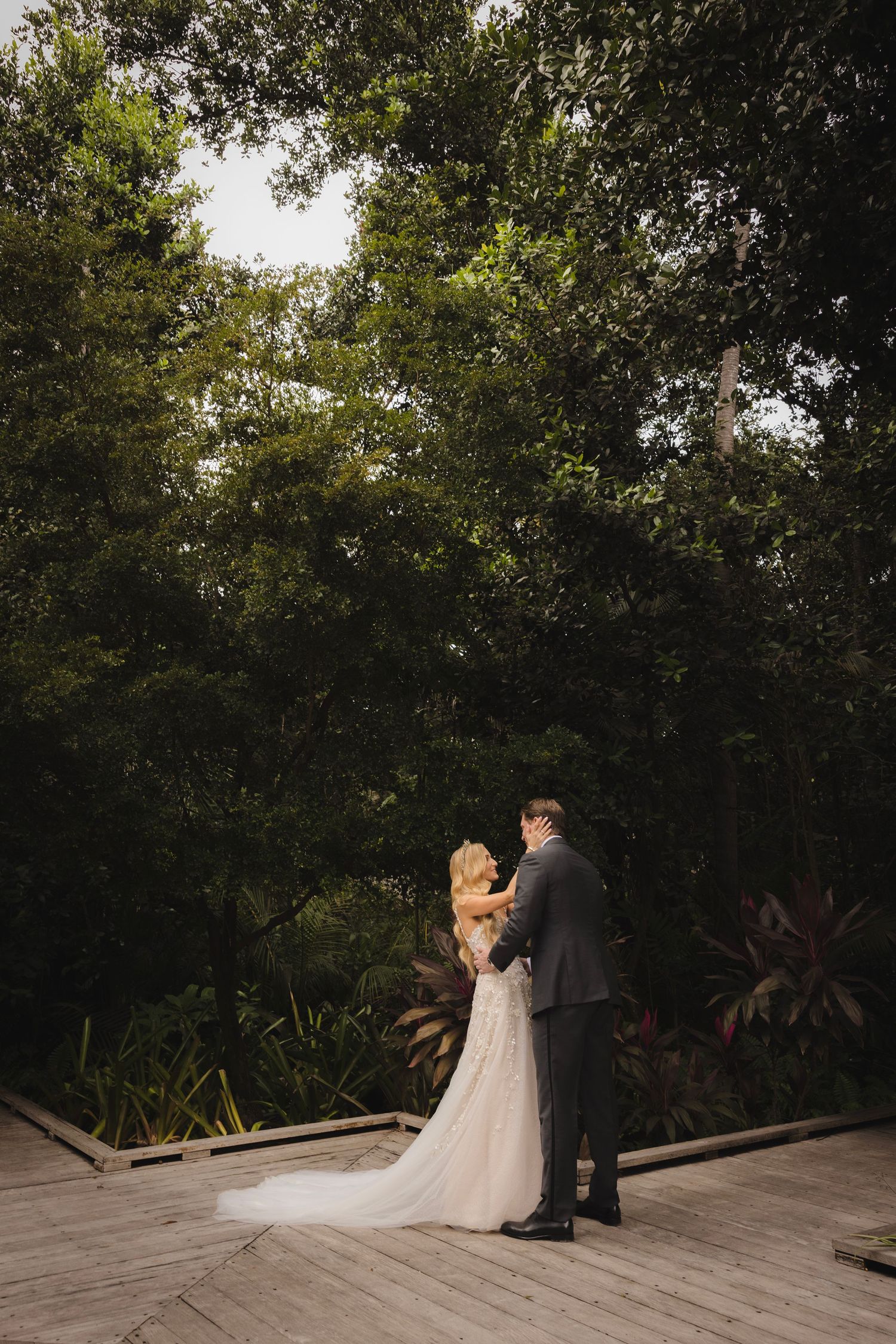A couple in wedding attire embrace on a wooden deck surrounded by lush greenery and trees.
