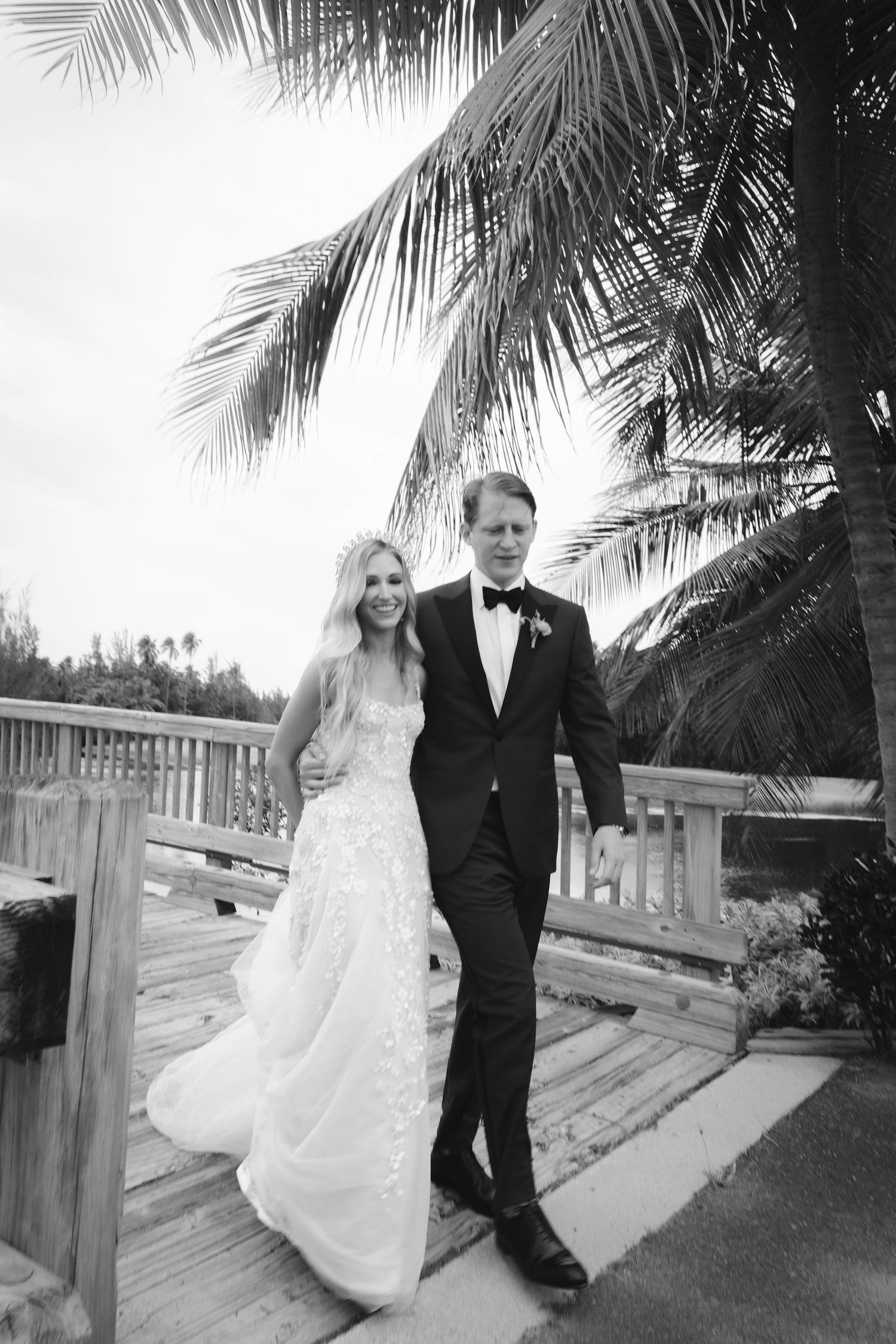 A wedding couple poses together under palm trees on a wooden boardwalk in black and white photography series.