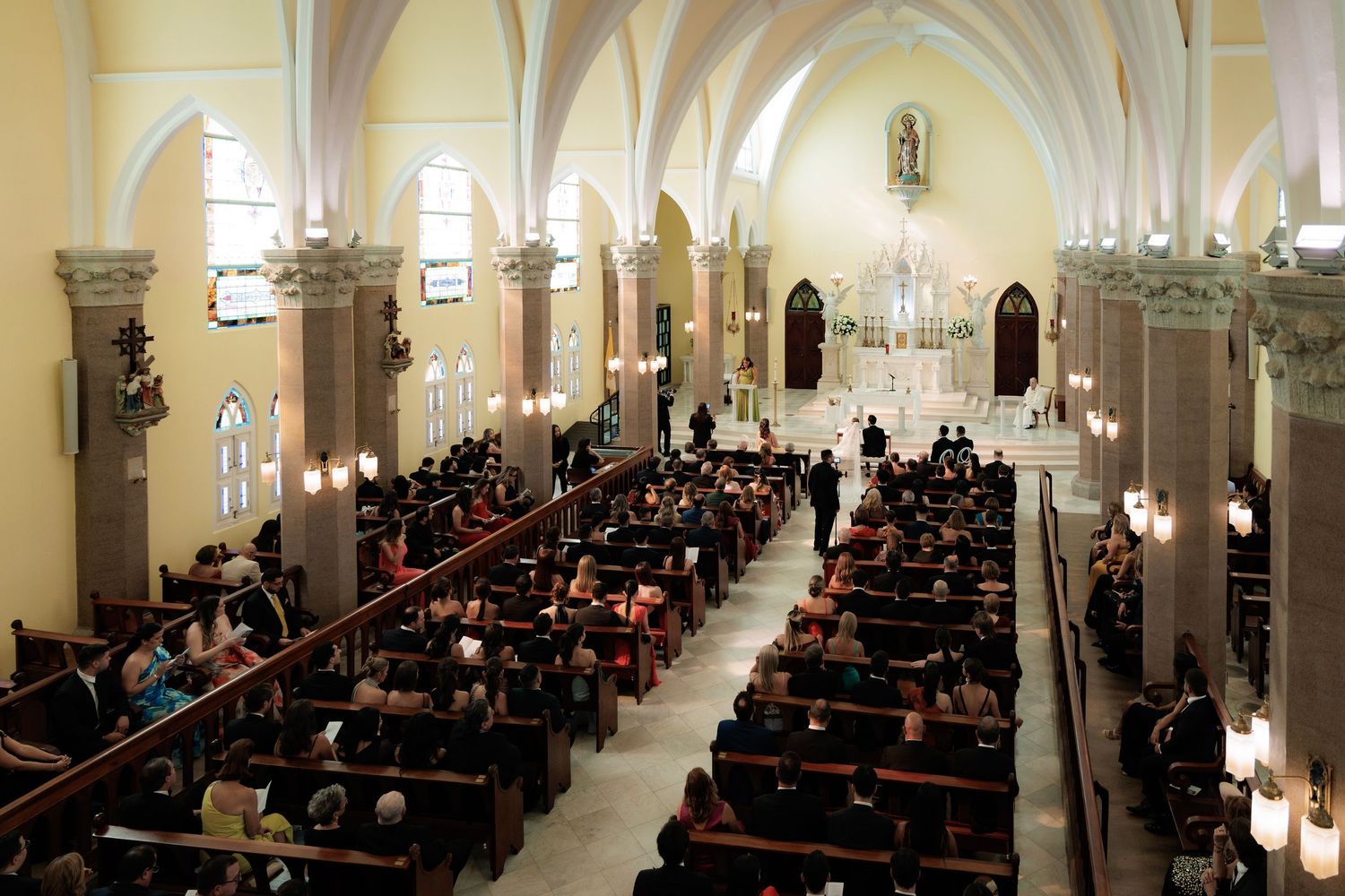 Worshippers gather inside a grand cathedral with white arched columns and wooden pews for an evening service.