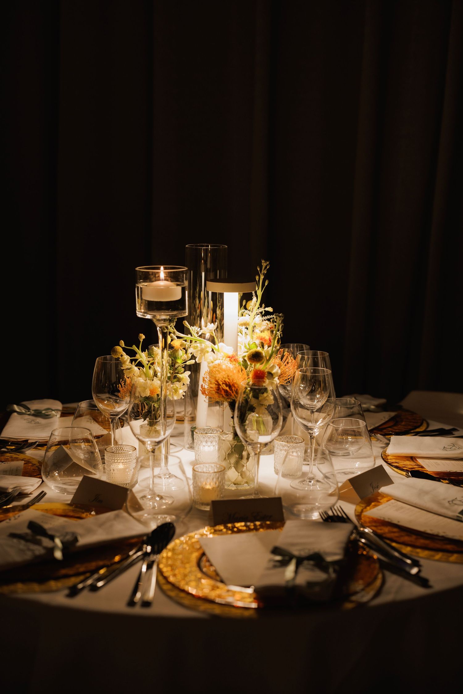 Elegant wedding reception table setting with candlelight, floral centerpieces and gold charger plates in dark ambiance.