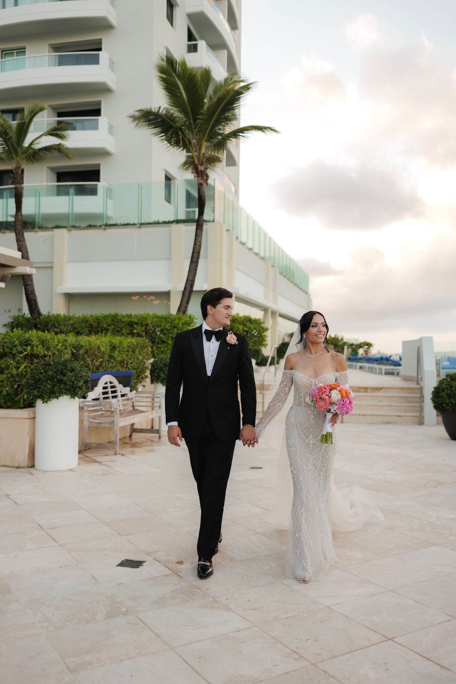 Newlyweds stroll on tropical resort terrace with palm trees and modern architecture.