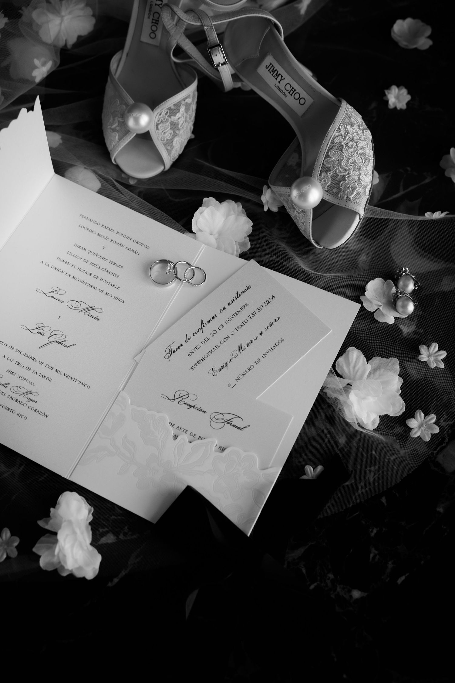 Black and white wedding invitation cards with white floral decorations and wedding rings arranged on a dark surface.