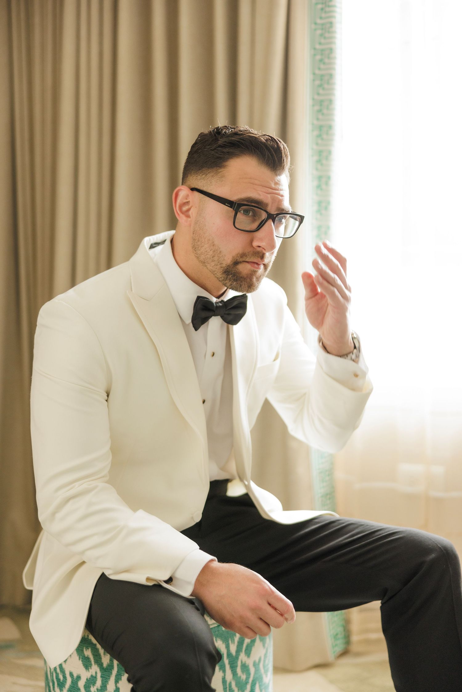 A groom in a white tuxedo jacket and black bow tie adjusts his cufflinks while getting ready.