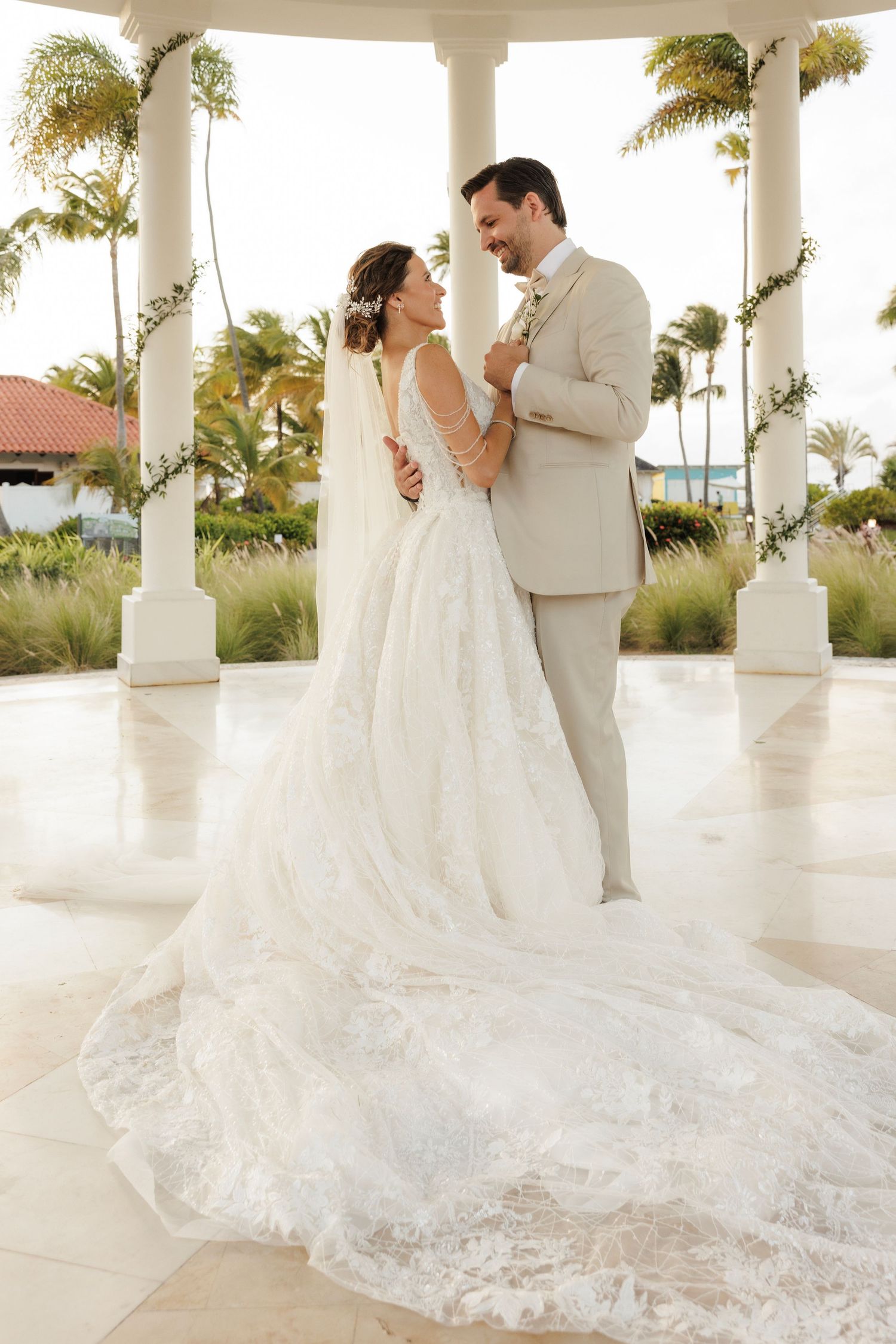 A couple shares a romantic moment under a white columned gazebo surrounded by palm trees at sunset for their wedding.