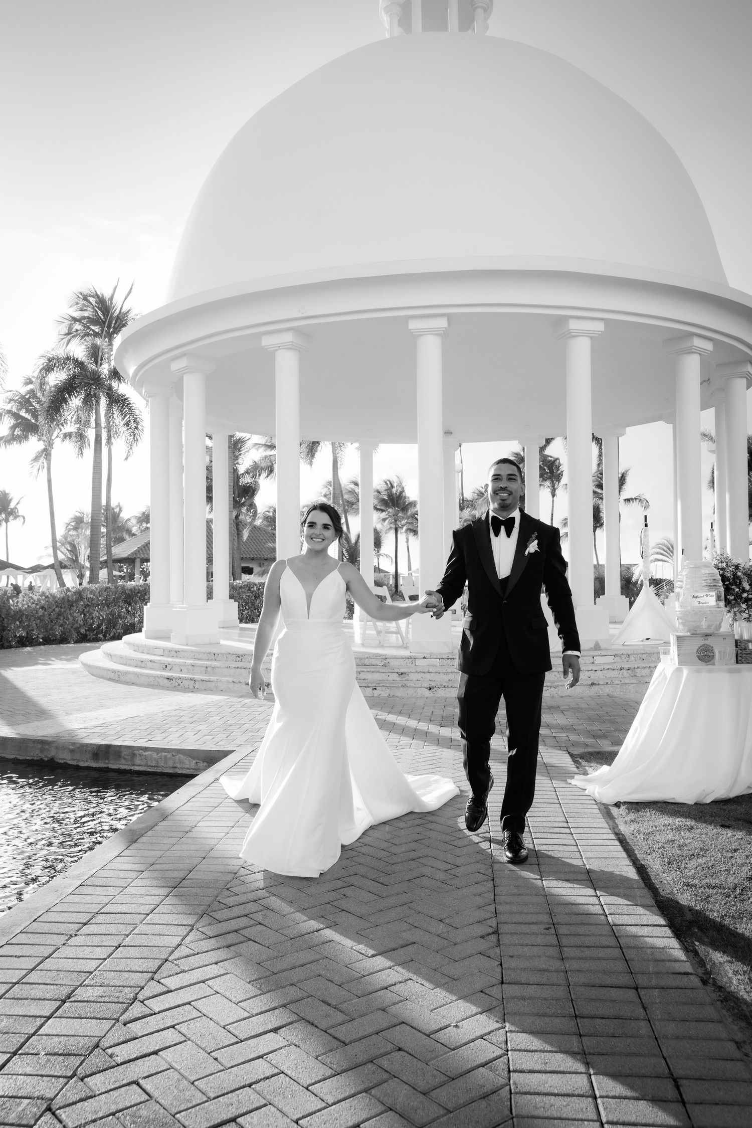 Black and white wedding photo sequence showing couple walking under elegant white archways by tropical palm trees.