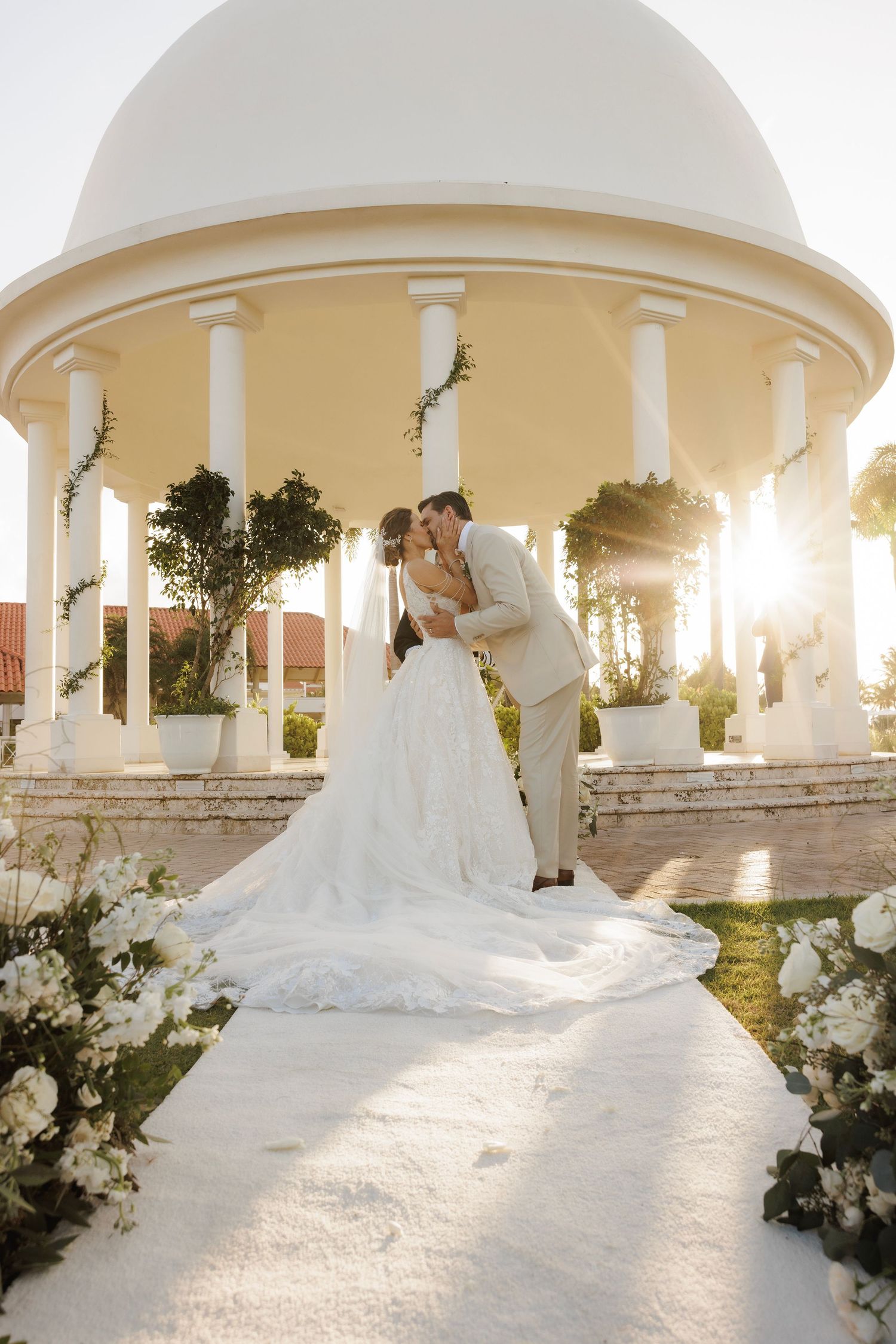 A bride in a flowing white wedding gown with a dramatic long train poses in an elegant columned outdoor pavilion at sunset.