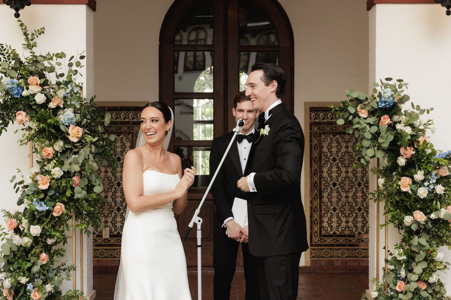 A wedding ceremony takes place in front of elegant wooden doors with floral arch arrangements in peach and white.
