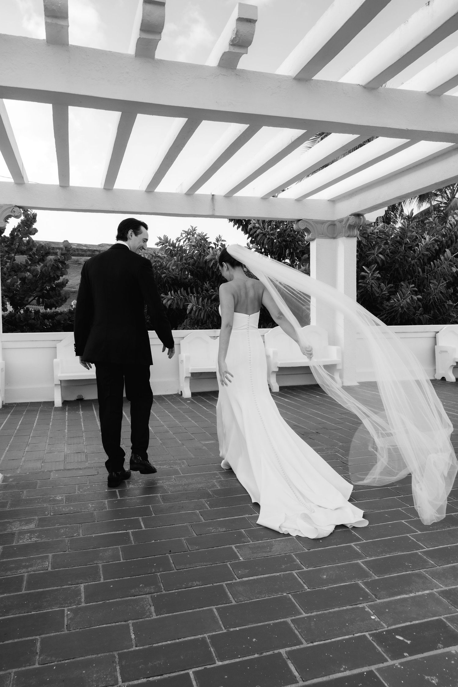 Wedding couple walking under a white pergola on a stone patio creating flowing movement with the dress.