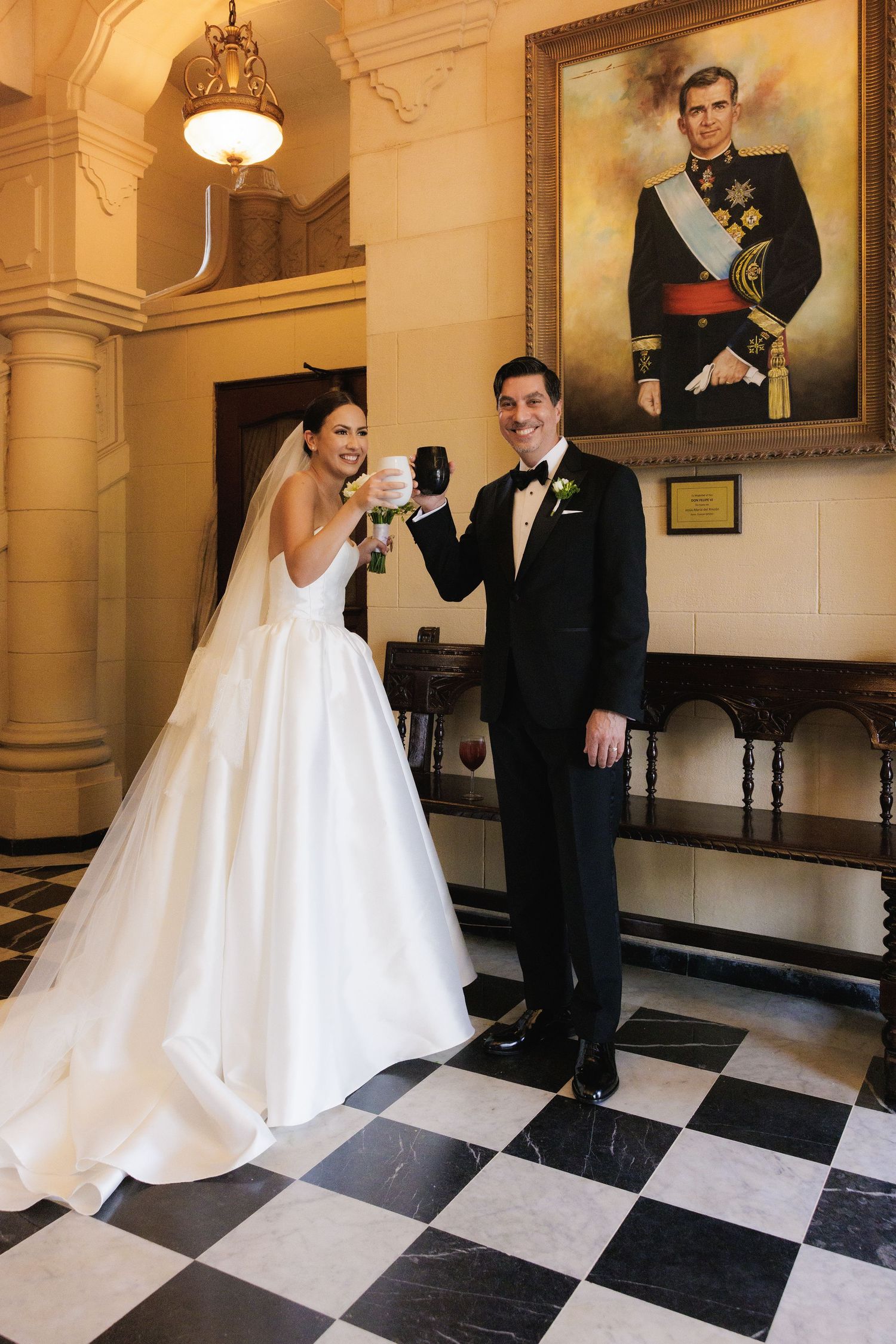 A wedding couple dances on a black and white checkered floor in an elegant ballroom with portraits on the wall.