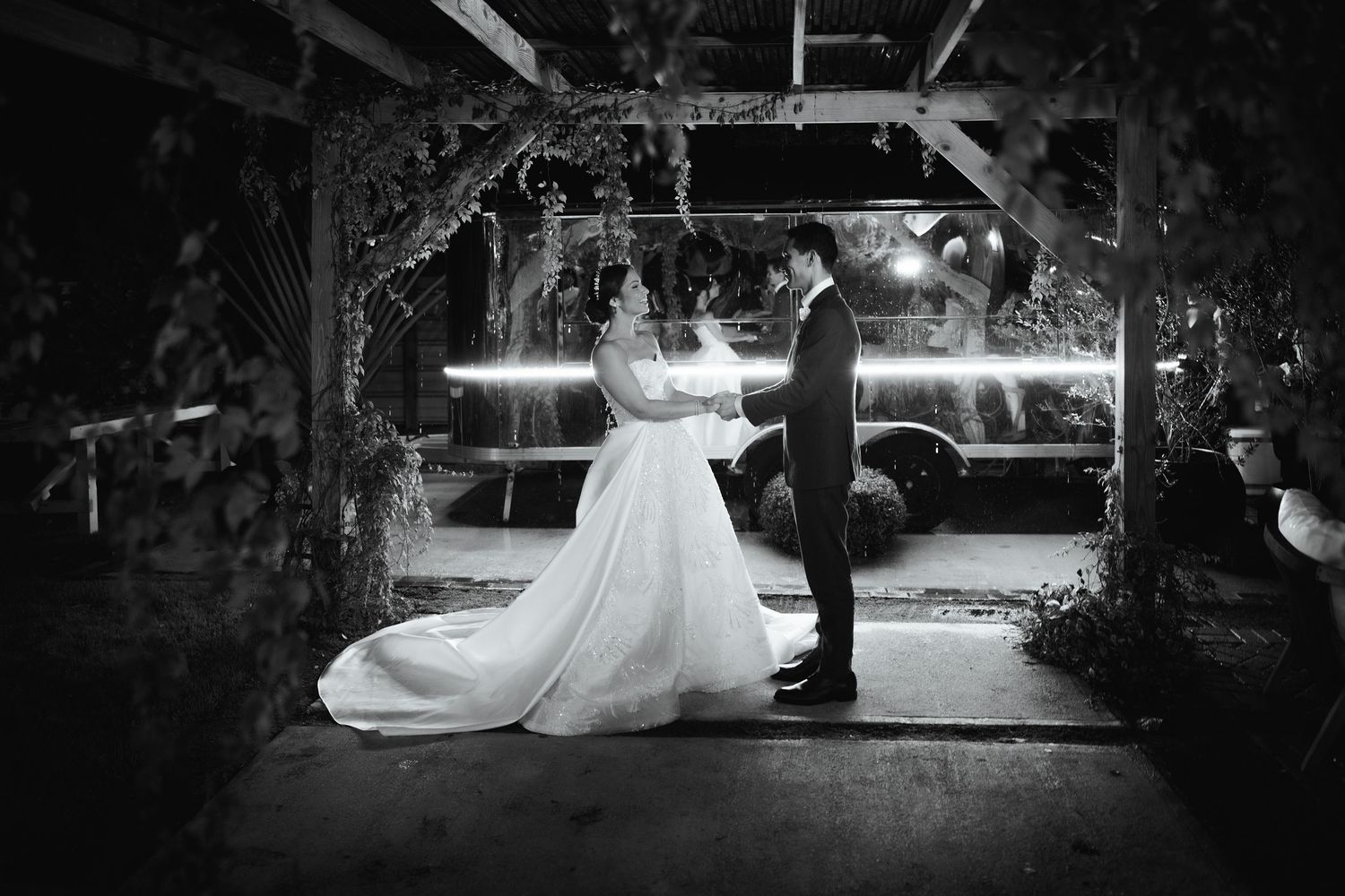 A romantic black and white wedding photo showing a couple dancing under a decorated arch at night.