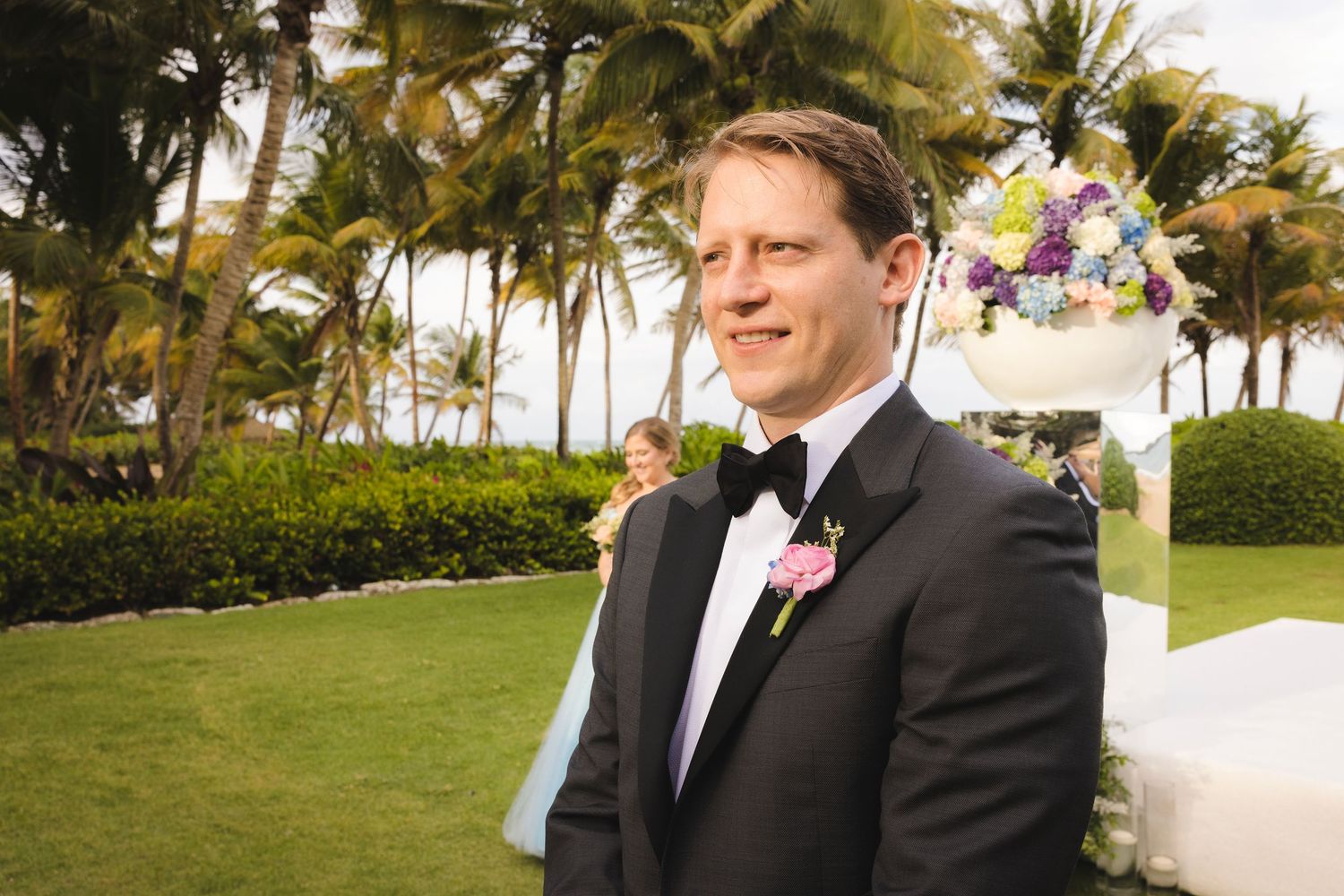 A person in a black tuxedo with pink boutonniere stands in front of palm trees at an outdoor wedding ceremony.