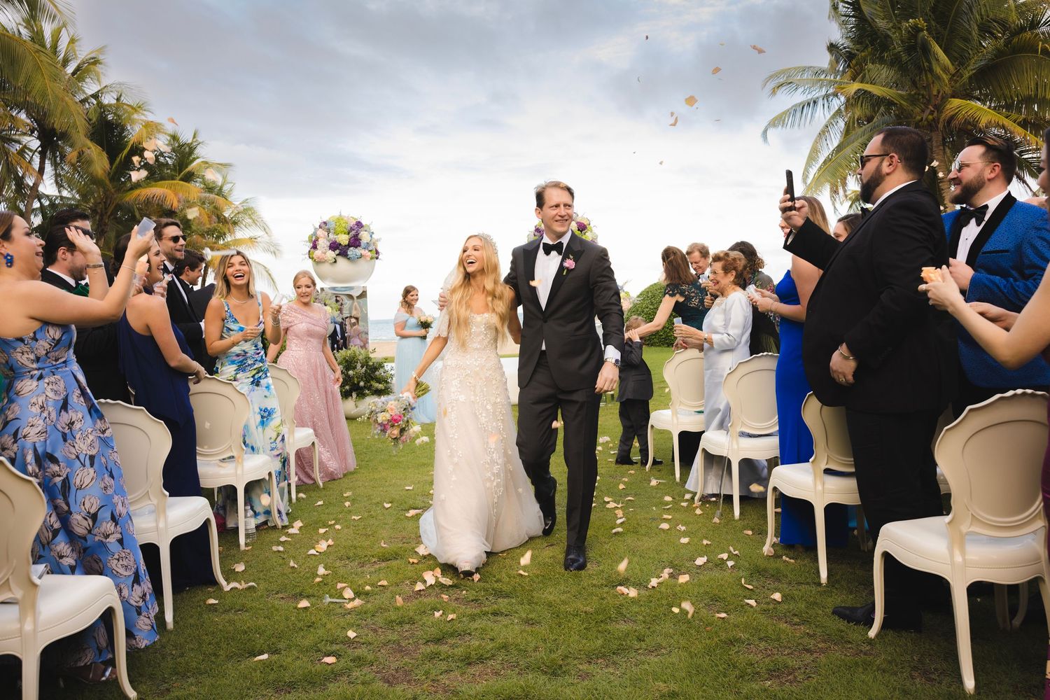 Wedding couple walks down floral aisle lined with white chairs and guests throwing petals at tropical beachfront venue.