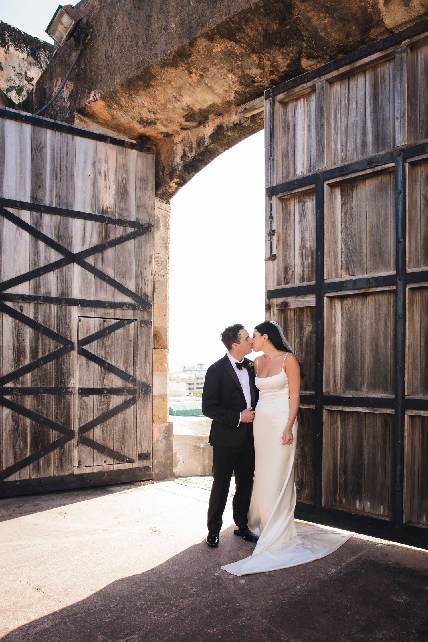 Newlyweds pose intimately by rustic wooden doors under a historic stone archway.
