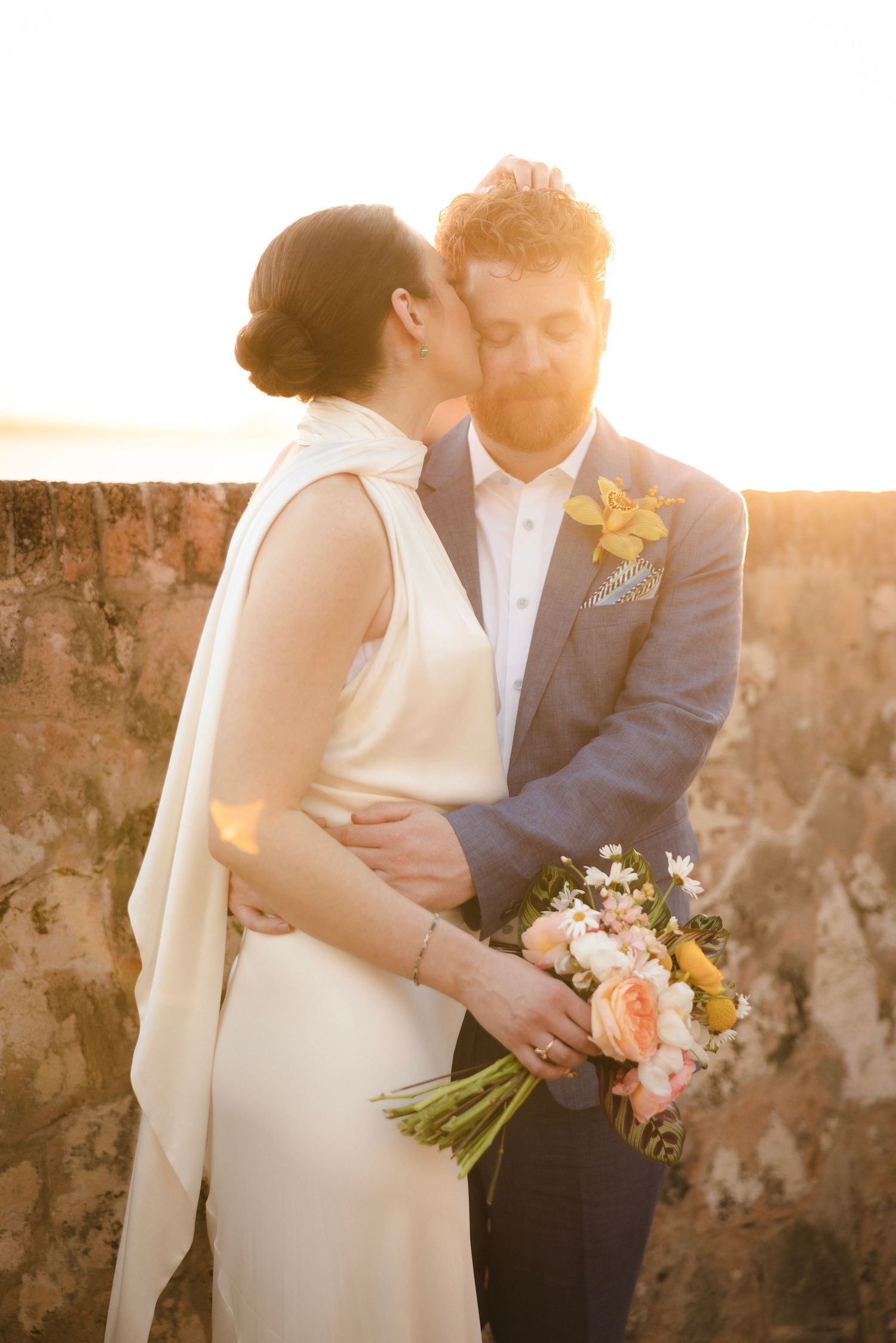 Romantic sunset wedding couple embracing while holding a peach and yellow bouquet against stone wall background.