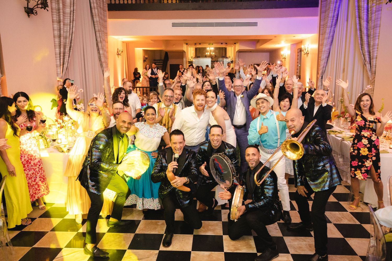 A vibrant wedding reception room with a dance floor full of party guests dancing and celebrating under purple and green lights.
