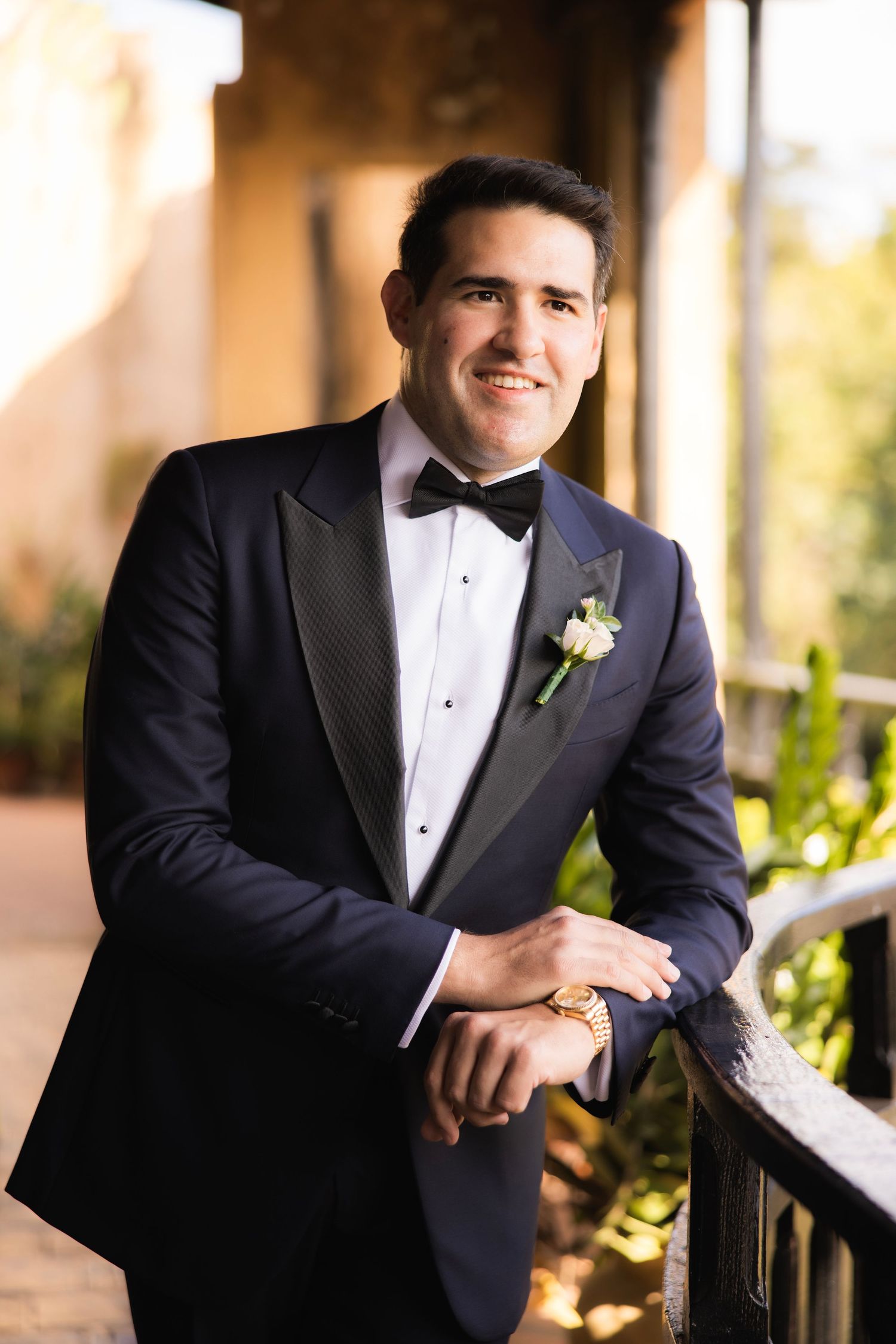 A groom in a classic black tuxedo and bowtie poses for formal wedding portraits in warm lighting.