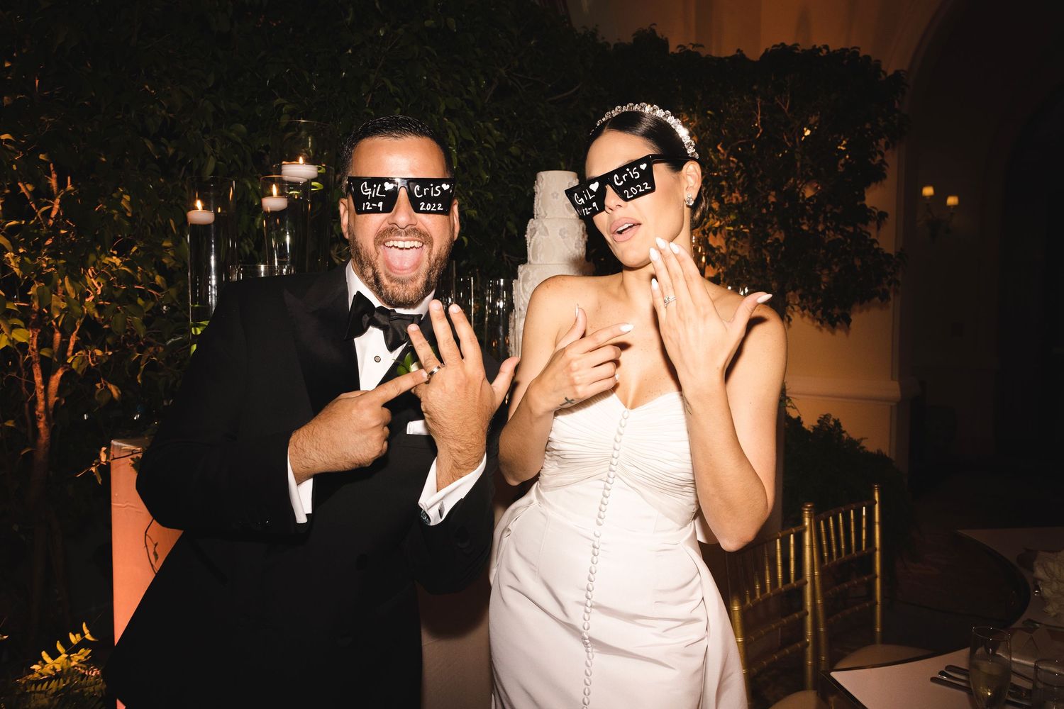 A couple in formal attire wearing sunglasses pose playfully for a wedding photo at an evening reception.