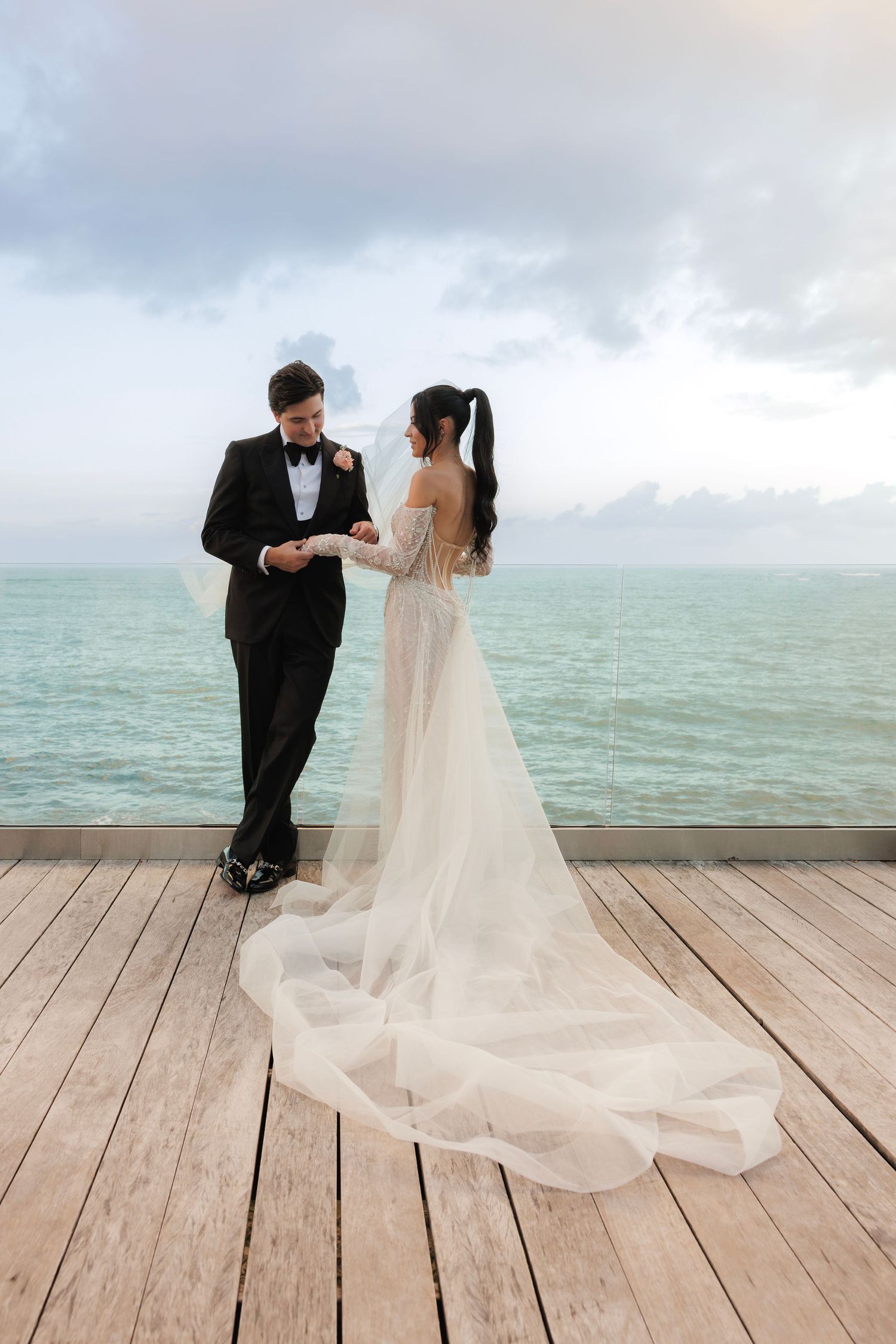 A couple stands together on a wooden pier overlooking turquoise waters during an overcast beach wedding ceremony.