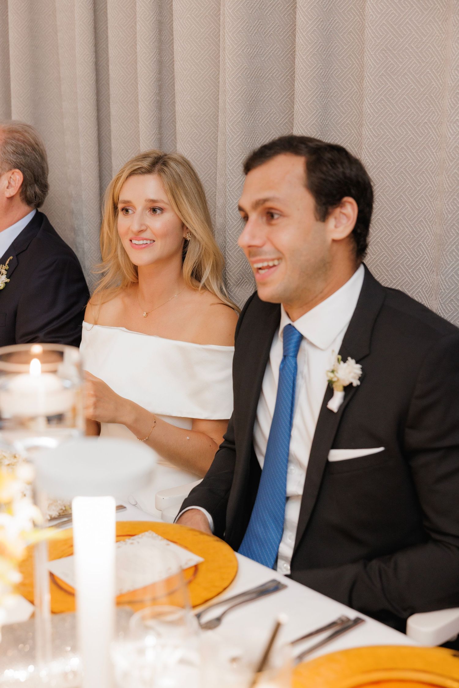 Wedding reception guests sit at a table with bright candlelight and formal place settings.