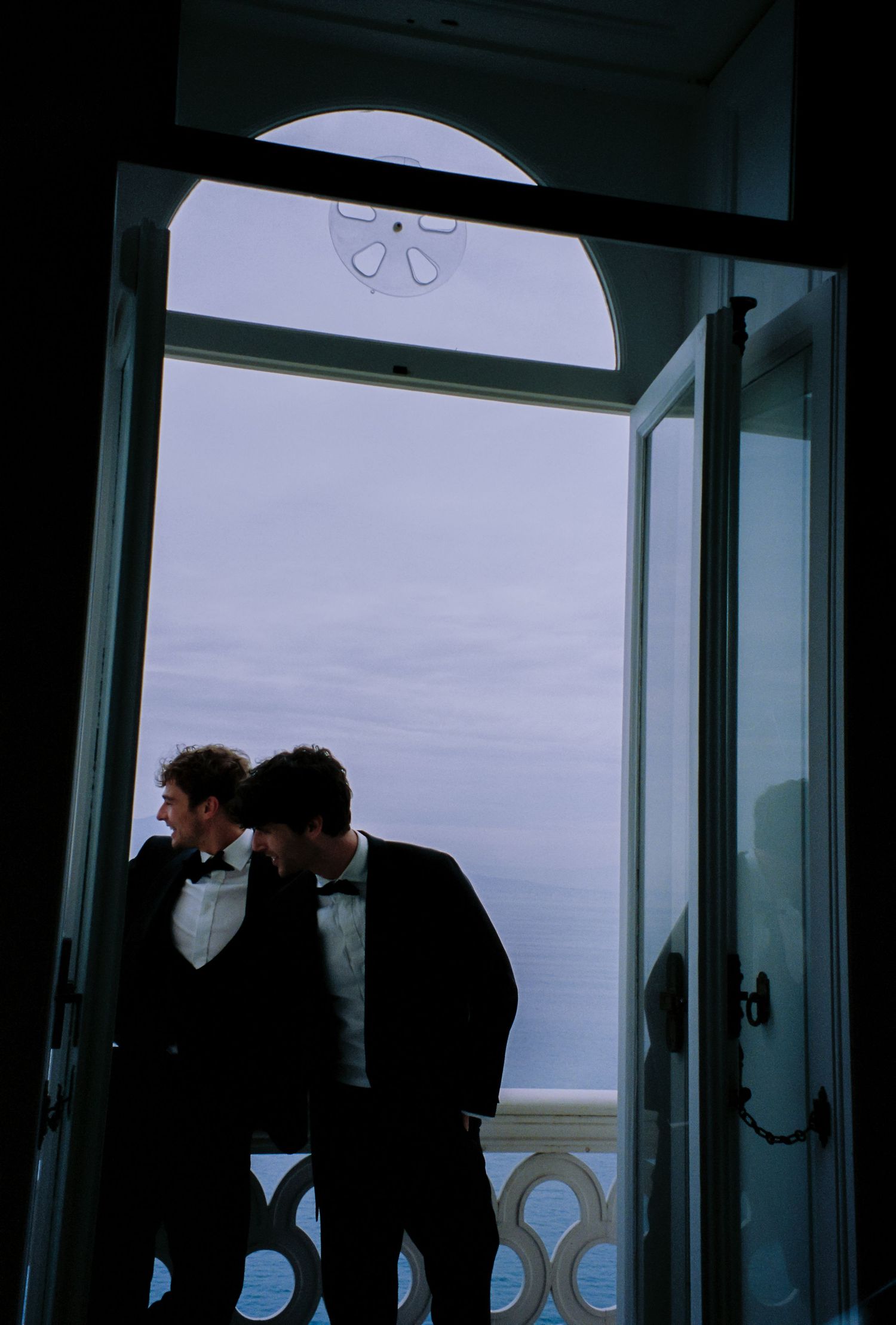 Two men in formal tuxedos stand in an open doorway overlooking the ocean at a seaside venue.