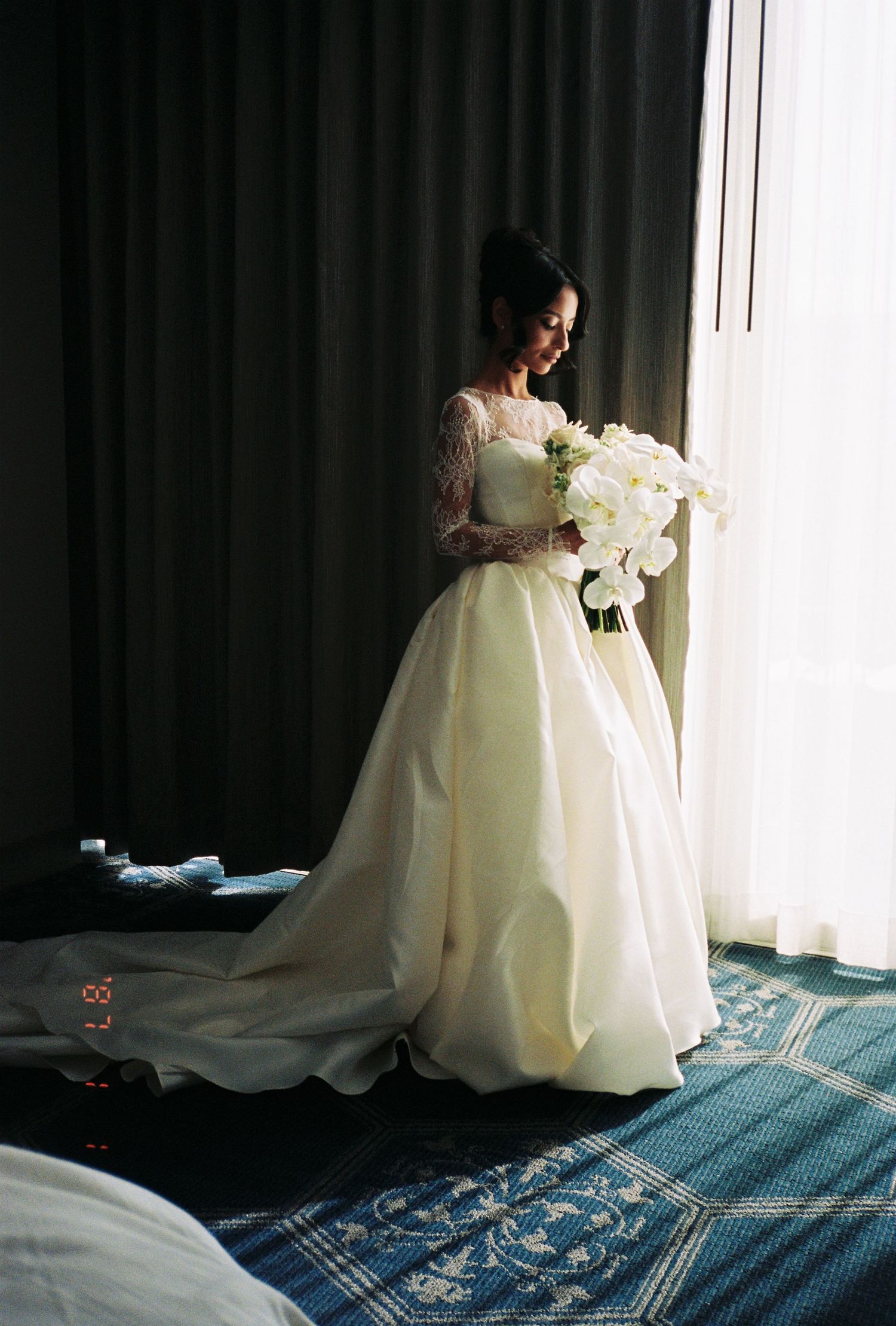 Bride in ivory ball gown with lace sleeves holding white bouquet standing by hotel window.