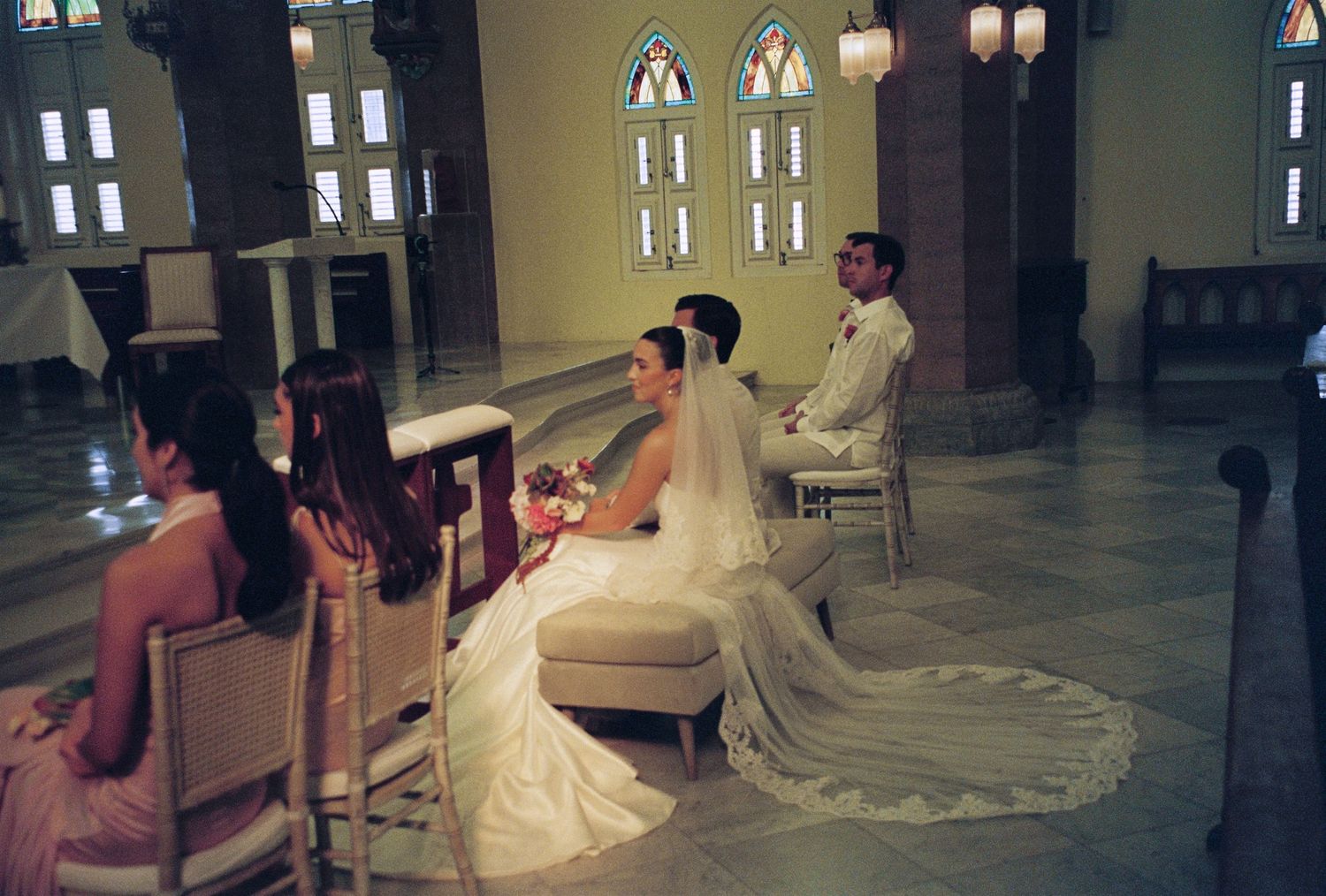 Bride and groom seated at church altar during wedding ceremony with bridesmaids nearby and stained glass windows.