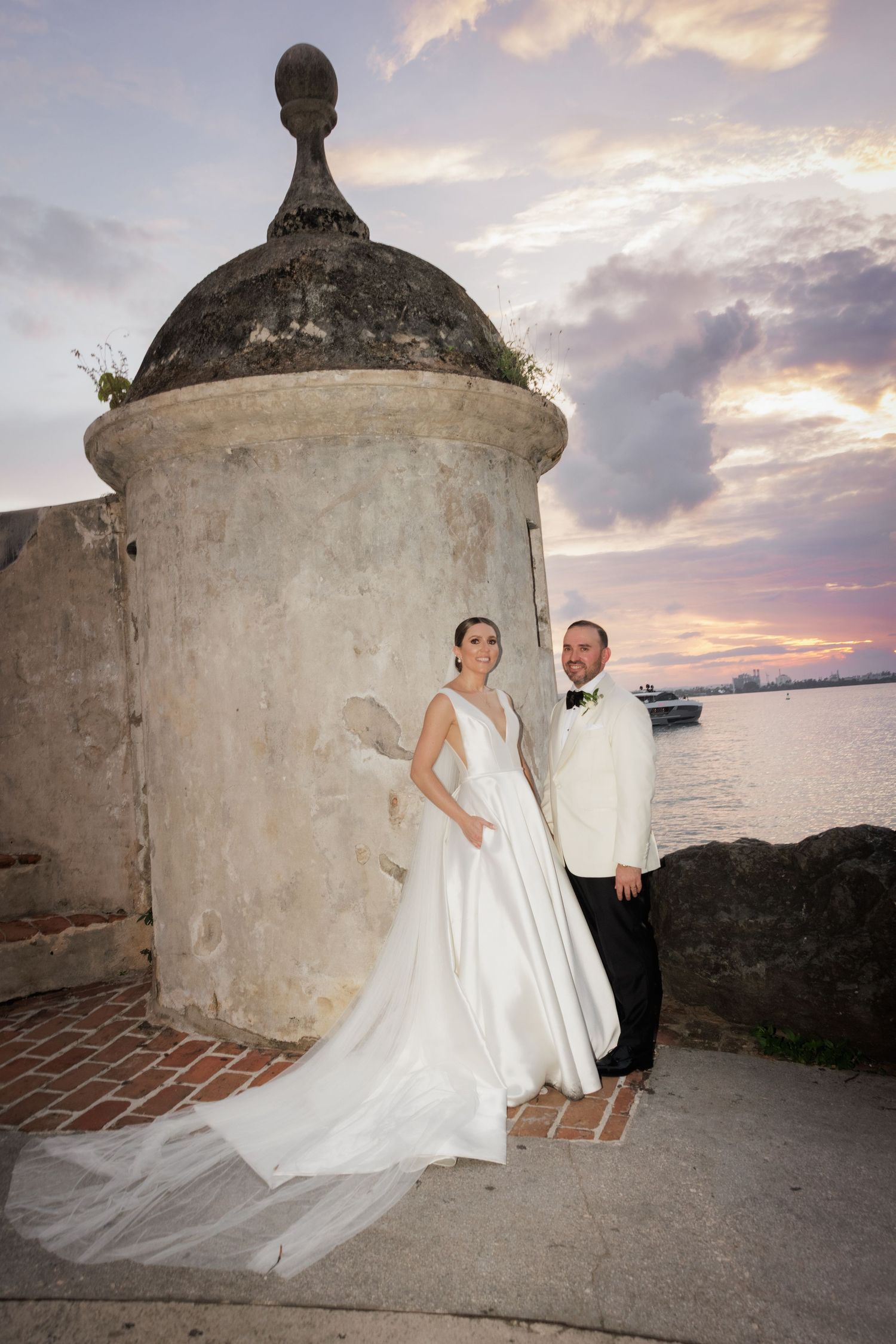 A wedding couple poses against stone walls at sunset with flowing white dress and dramatic cloudy sky in the background.