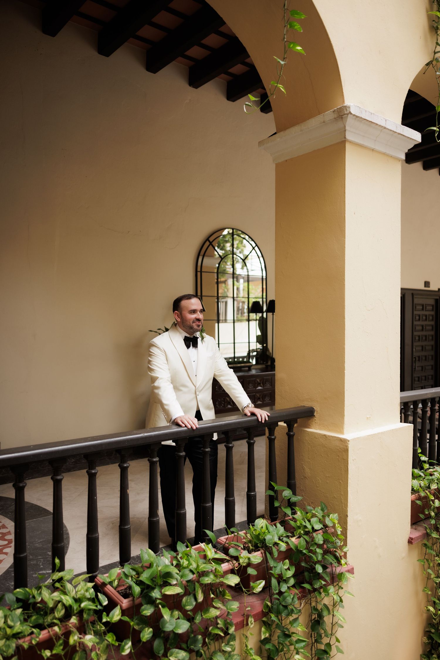A person in white stands on a Mediterranean-style balcony with arched columns and lush green foliage.