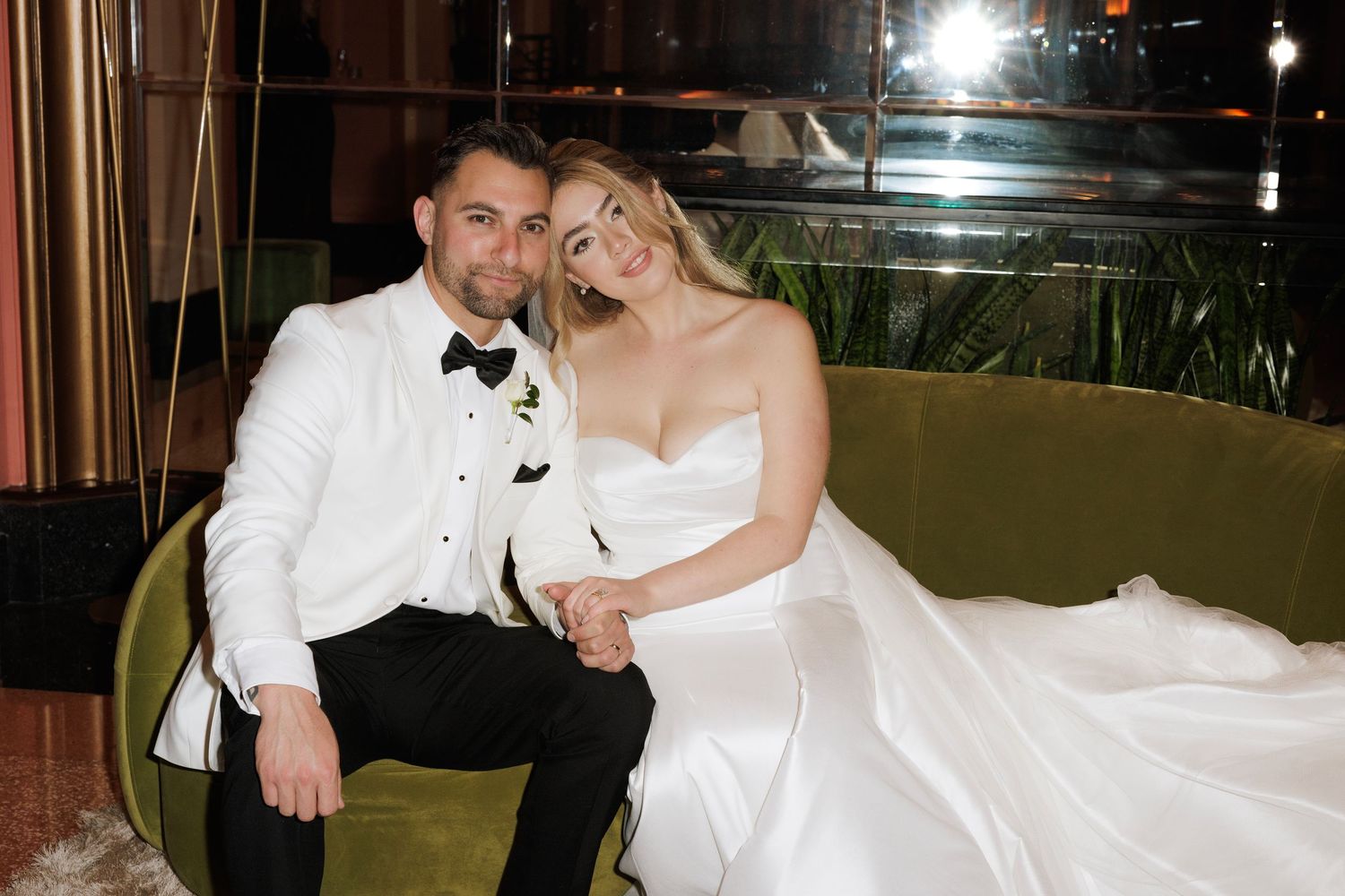 A couple in formal wedding attire sits together on a green velvet couch during their evening celebration.