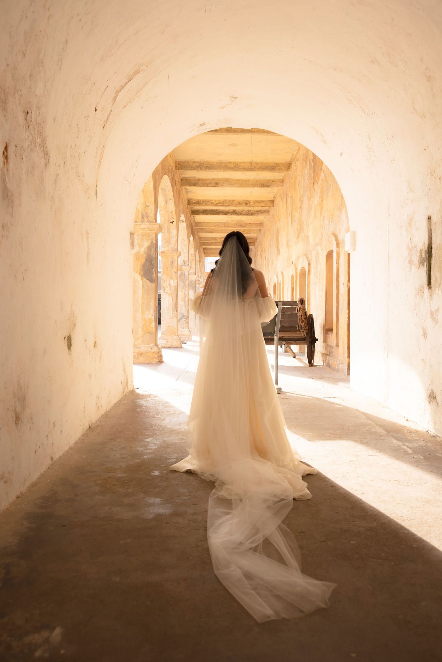 A flowing white wedding dress creates an ethereal silhouette against a sunlit stone corridor in a historic building.