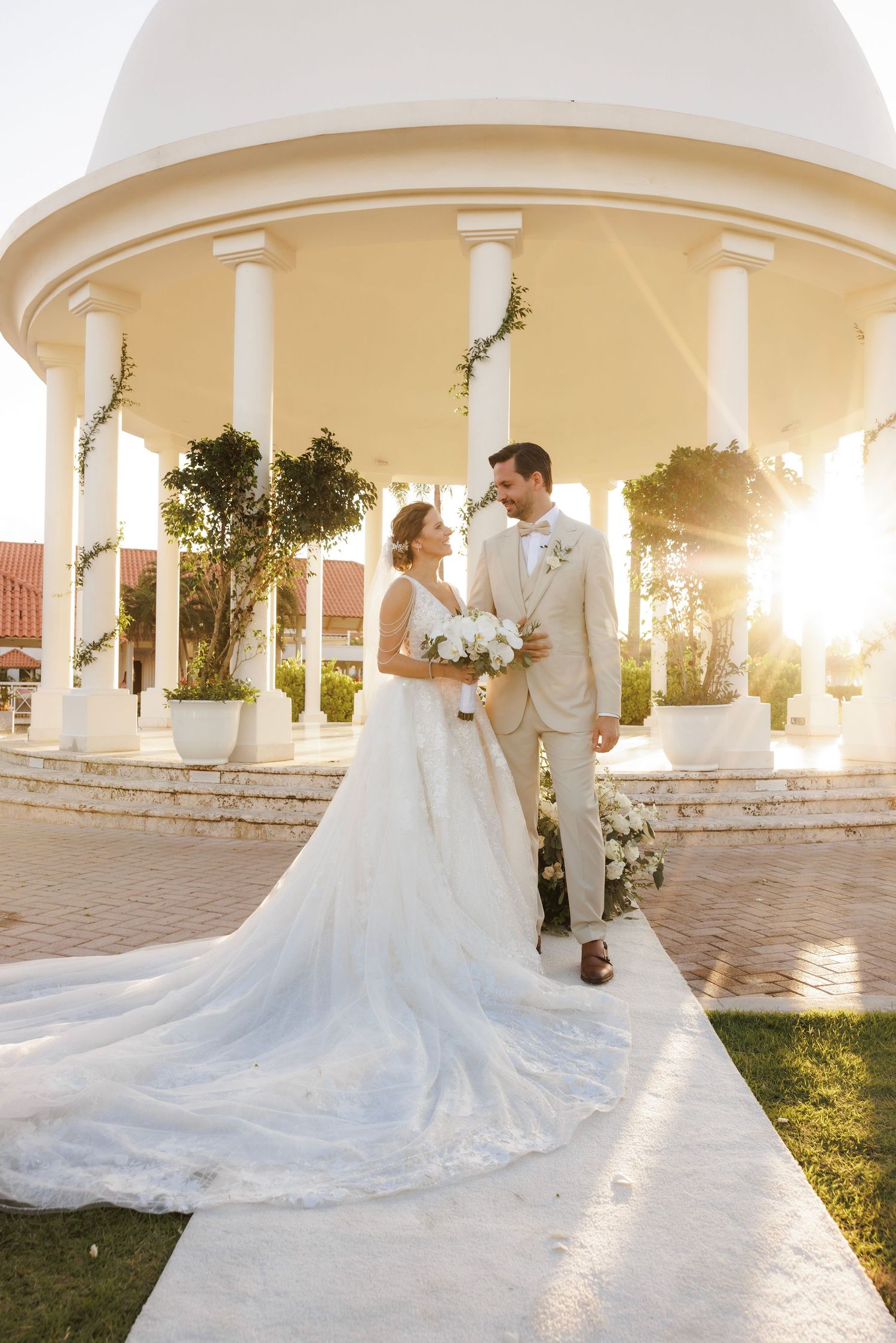 Golden sunset light illuminates archways and pillars as a couple in formal wedding attire embraces on a garden patio.
